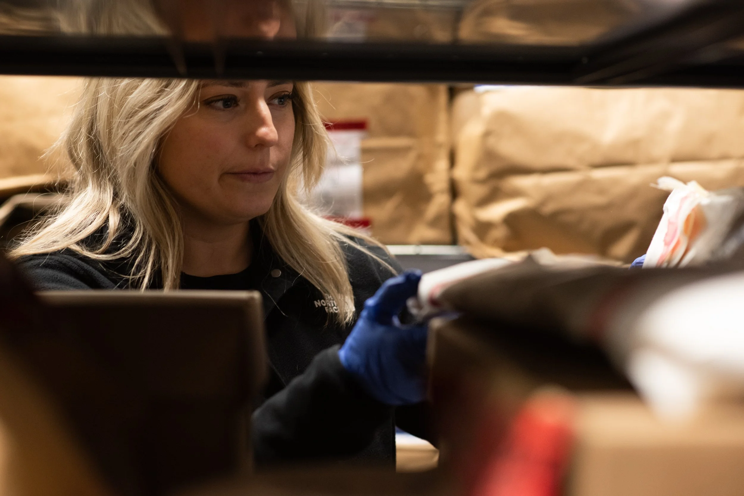 Woman with blonde hair wearing a black jacket and blue disposable gloves looking at items on a shelf inside a warehouse area.