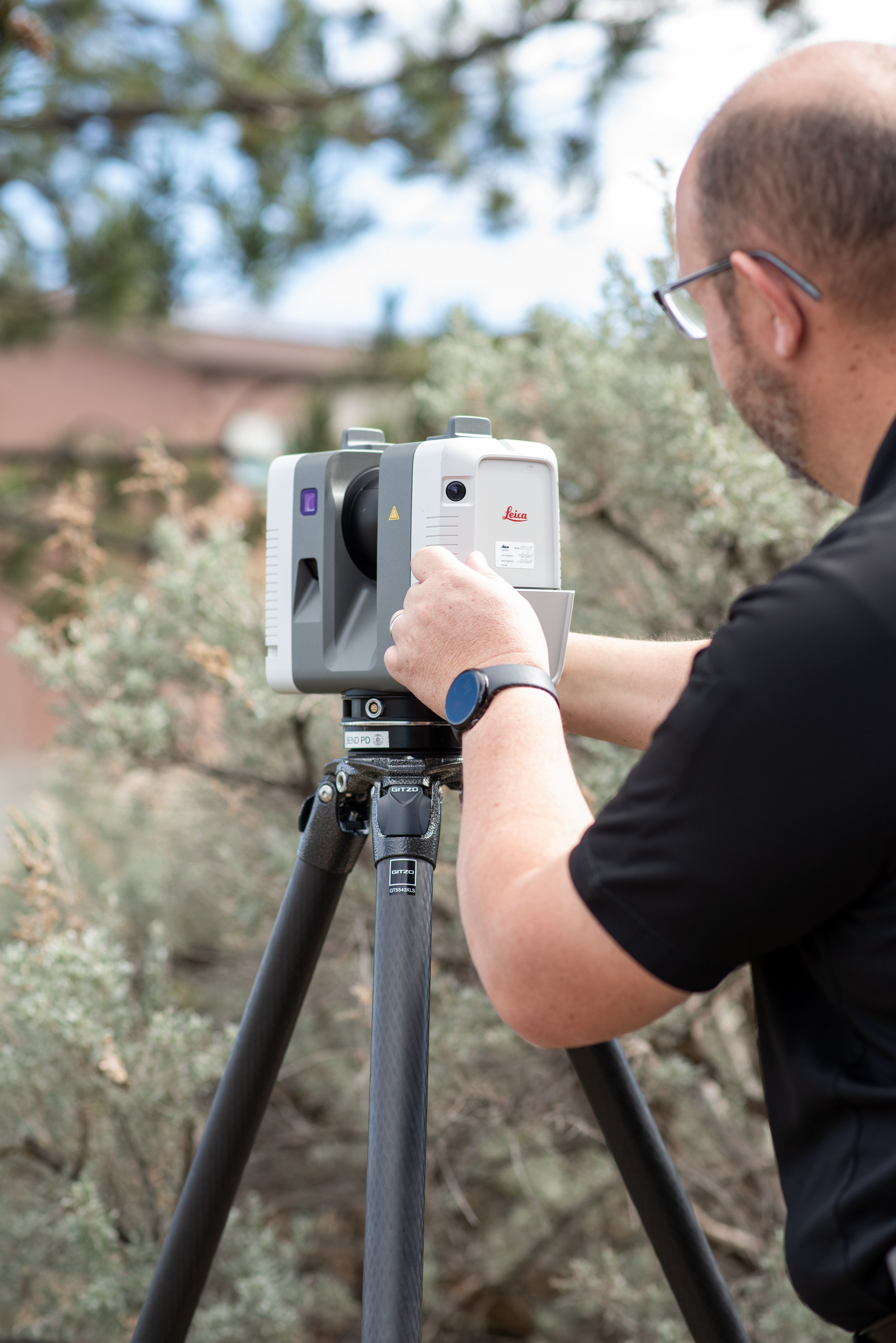 A man wearing glasses is facing away and changing setting on a Leica 360 degree camera mounted in a tripod with shrubs and a building in the background.