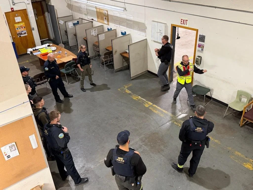 a small group of police officers inside a building are watching a person in a yellow safety vest give instructions to the group.