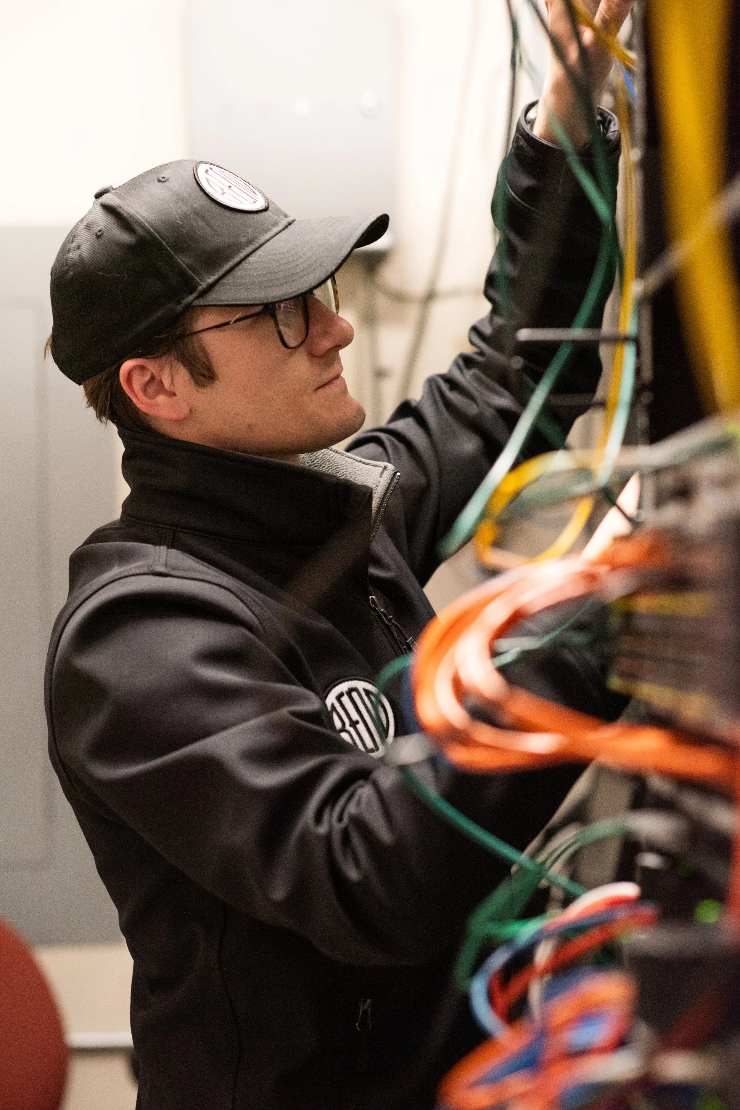 A man in a black baseball hat and a black jacket with the City of Bend logo is working on computer equipment surrounded by multi-colored wires.