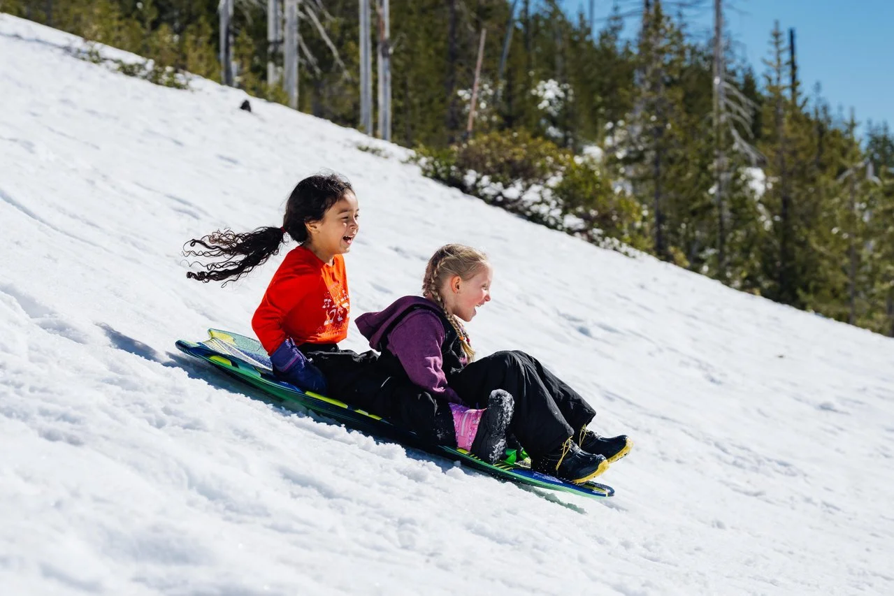 A girls with dark curly hair wearing and orange shirt and a girl with blong braids wearing a purple winter coat are smiling and laughing as they sled down a steep snow-covered hill on a sunny day with trees in the background.