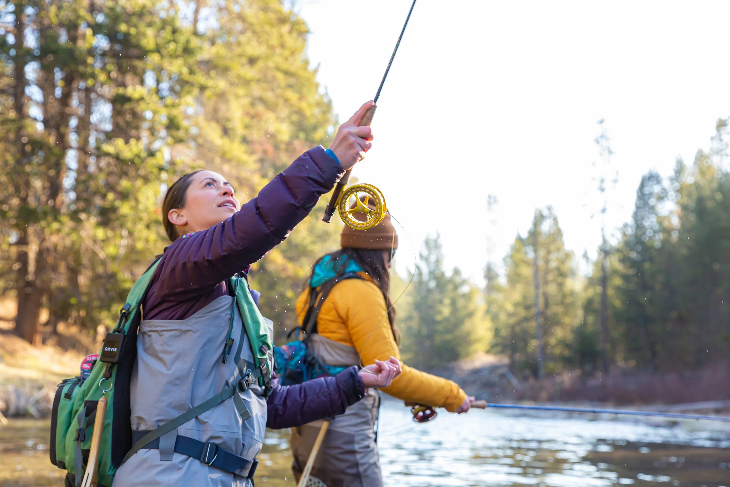Two women are standing outdoors in a river wearing fly fishing waders. One woman is wearing a purple jacket and casting a yellow fly rod. The other woman is wearing a yellow jacket and a brown knit hat and she is reeling in her fishing line.