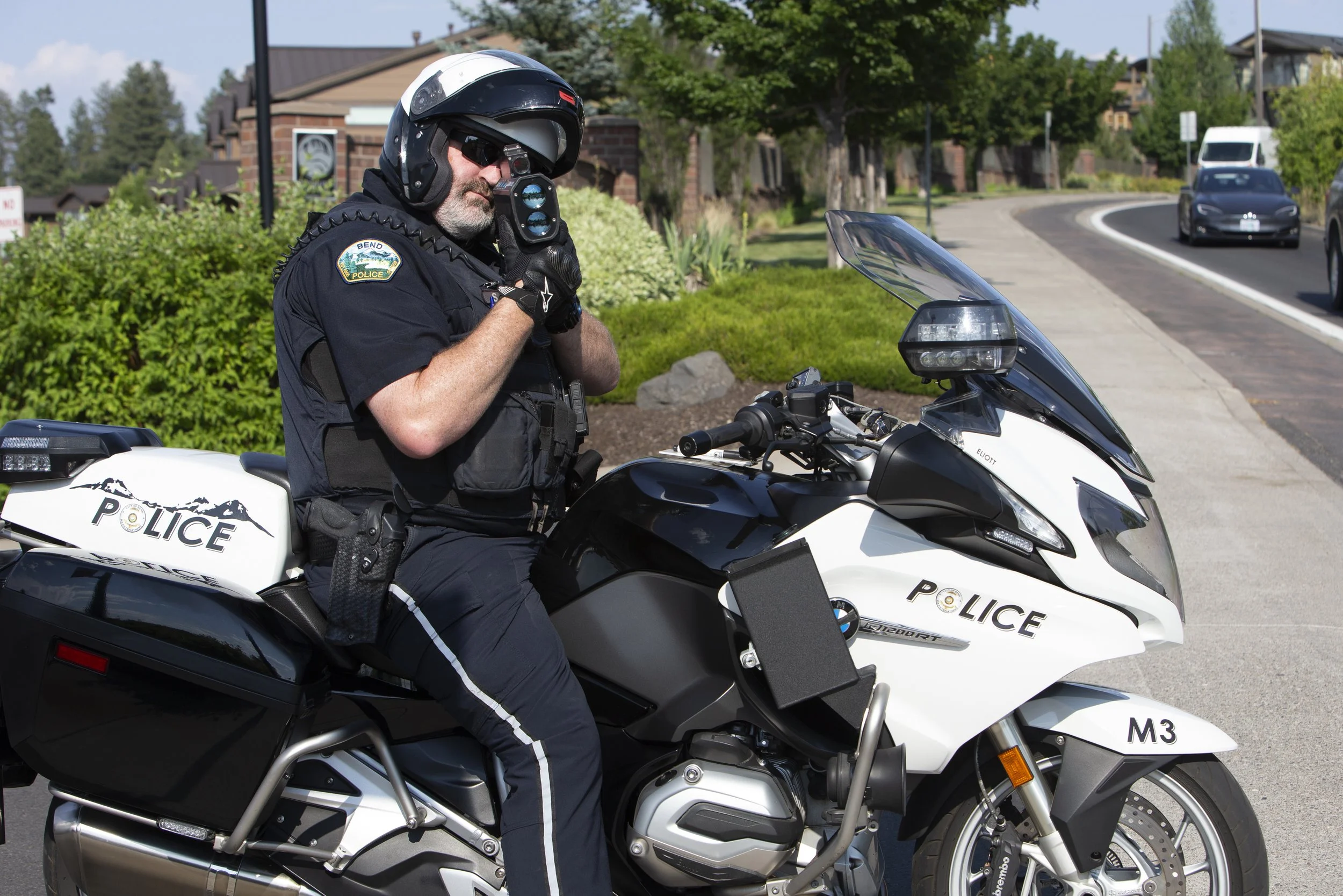 A police officer in uniform sitting on a police motorcycle on the side of city street, holding a LiDAR device to his face to measure vehicle speeds.