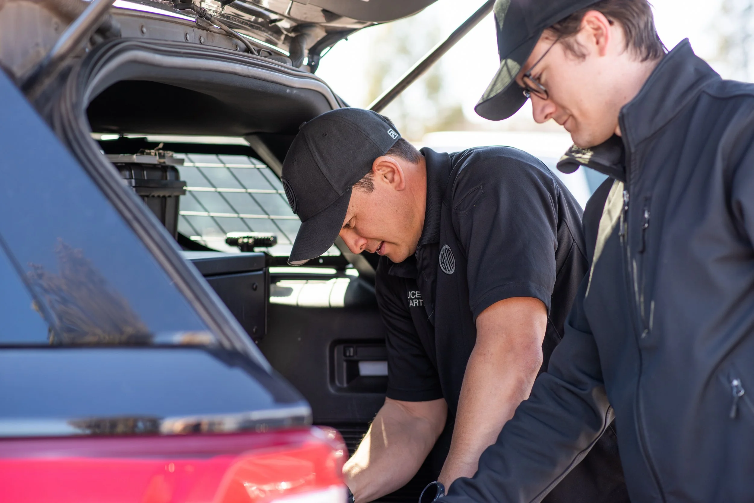 Two men in black clothing inspecting the trunk of a police vehicle outdoors.
