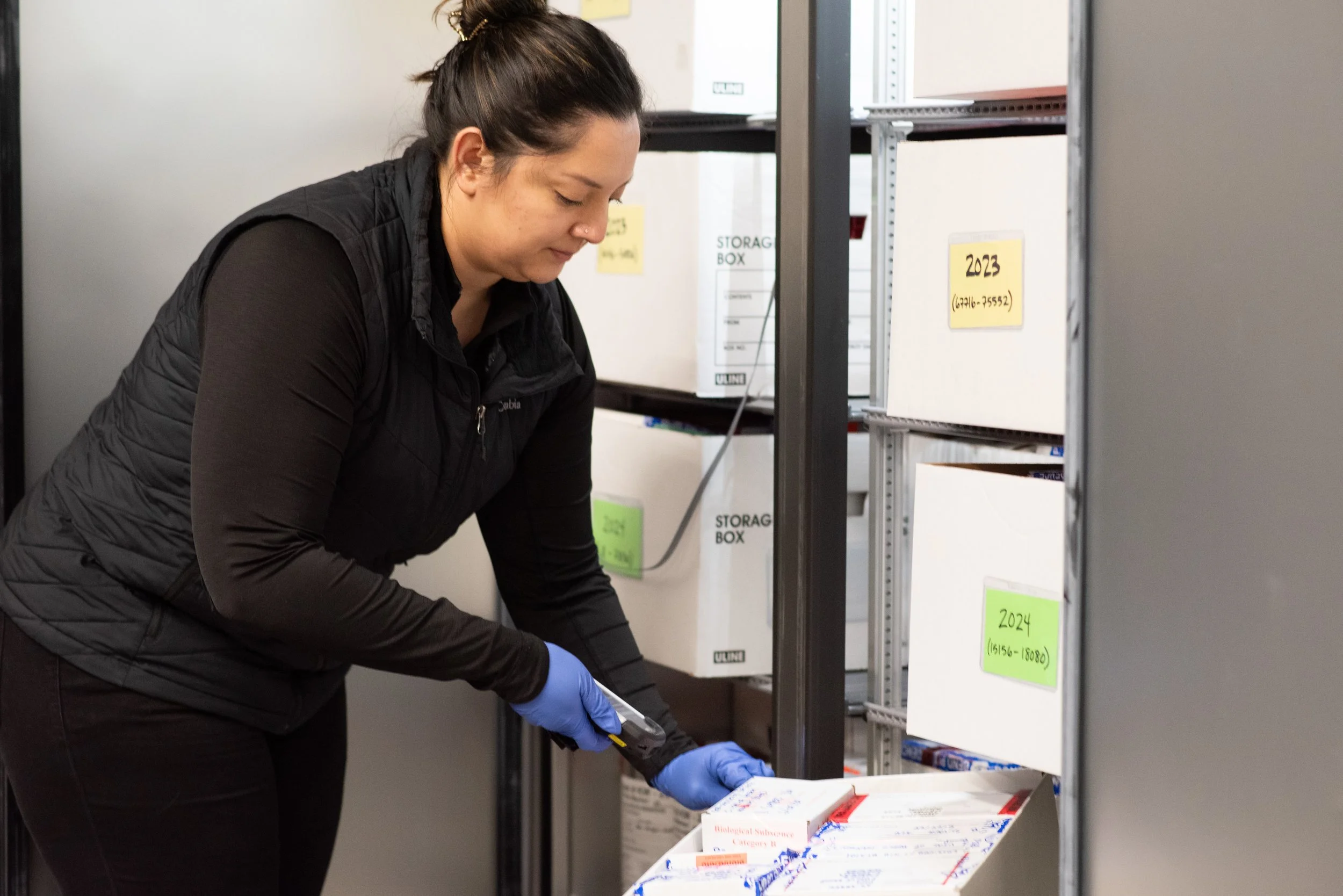 A woman wearing black clothing and blue disposable gloves is using a handheld scanner to scan evidence packaging inside a refrigerator.