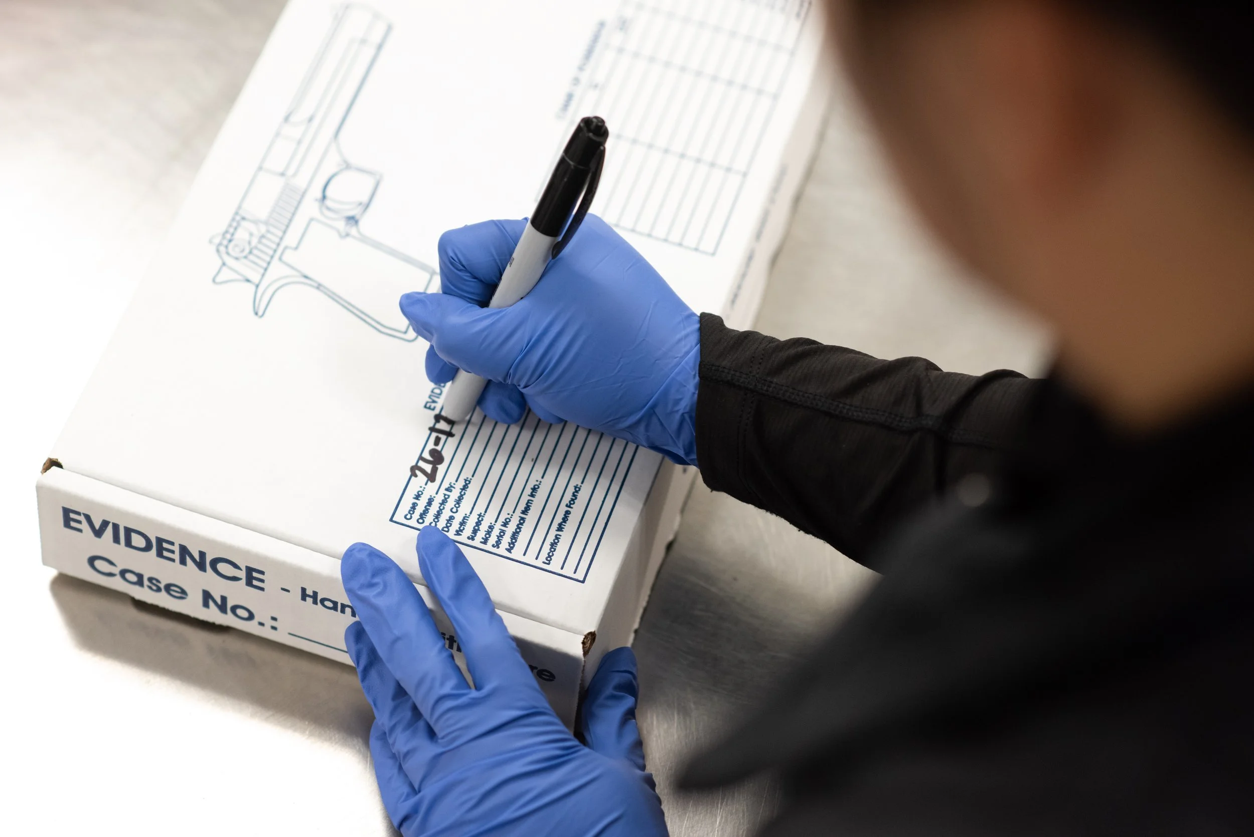 A person is writing on a box used to store pistols for evidence. The person is wearing blue disposable gloves and writing with a black marker. The box is on a stainless-steel table.
