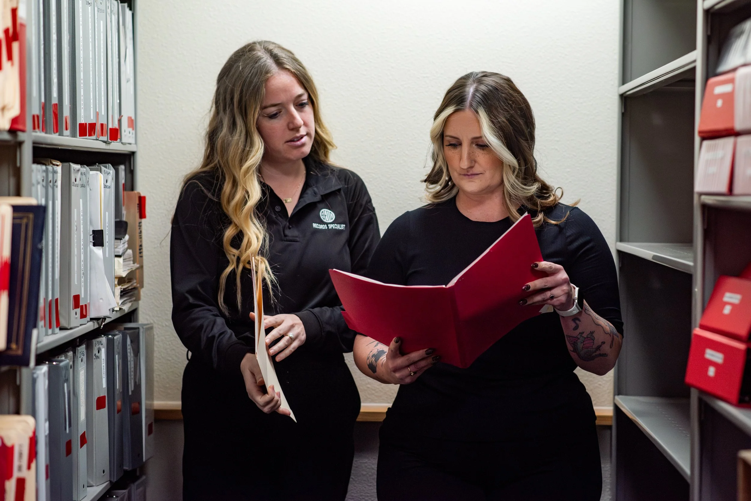 Two women are standing together between rows of file storage. They are holding filing folders. One folder is red, it is open and the women are looking at the contents together. The women are wearing black clothing.