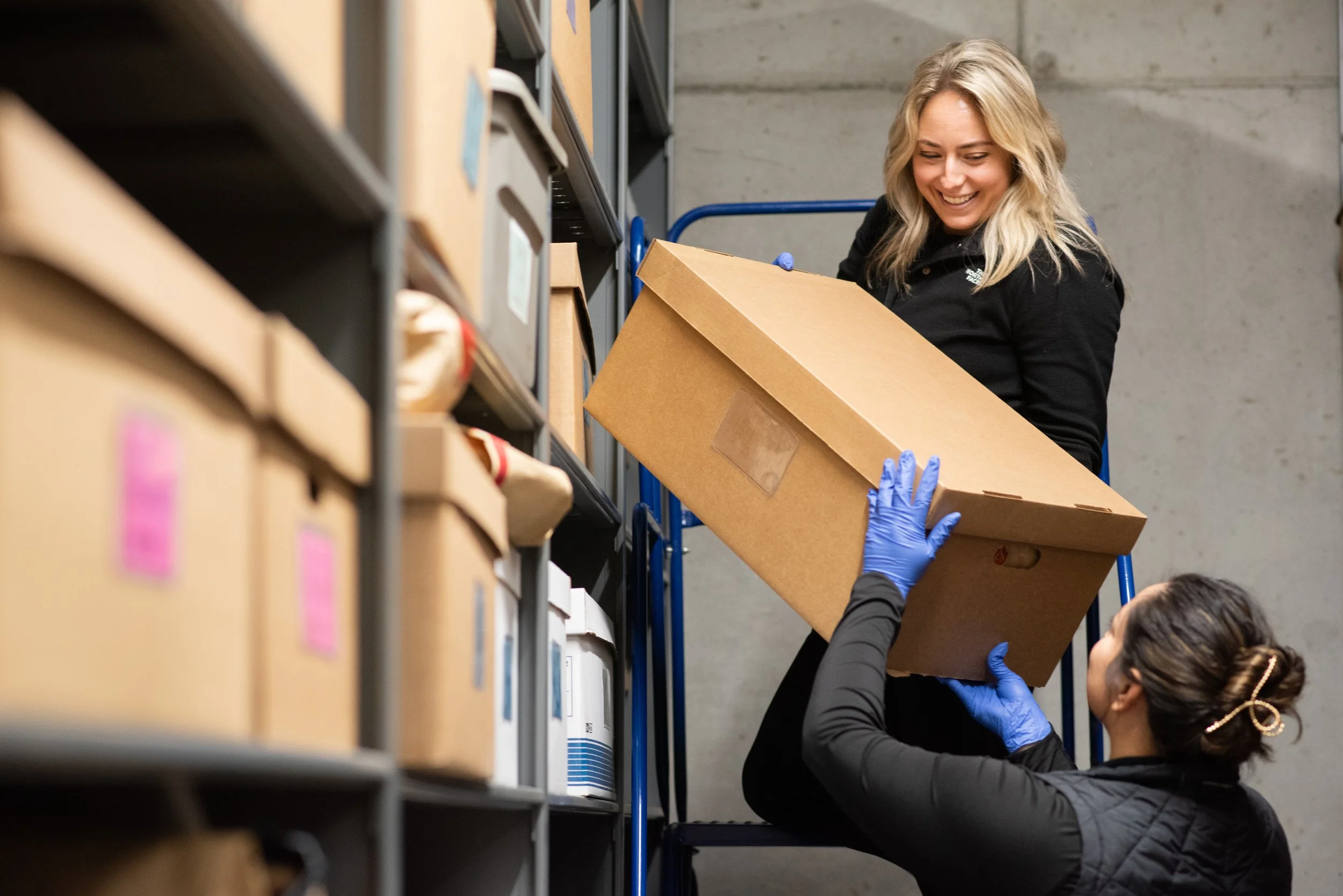 Two women are moving a large brown cardboard box in a storage space. One woman is standing on a blue ladder. She is smiling and handing the box to another woman who is standing on the ground. Both women are wearing black clothing and blue disposable 