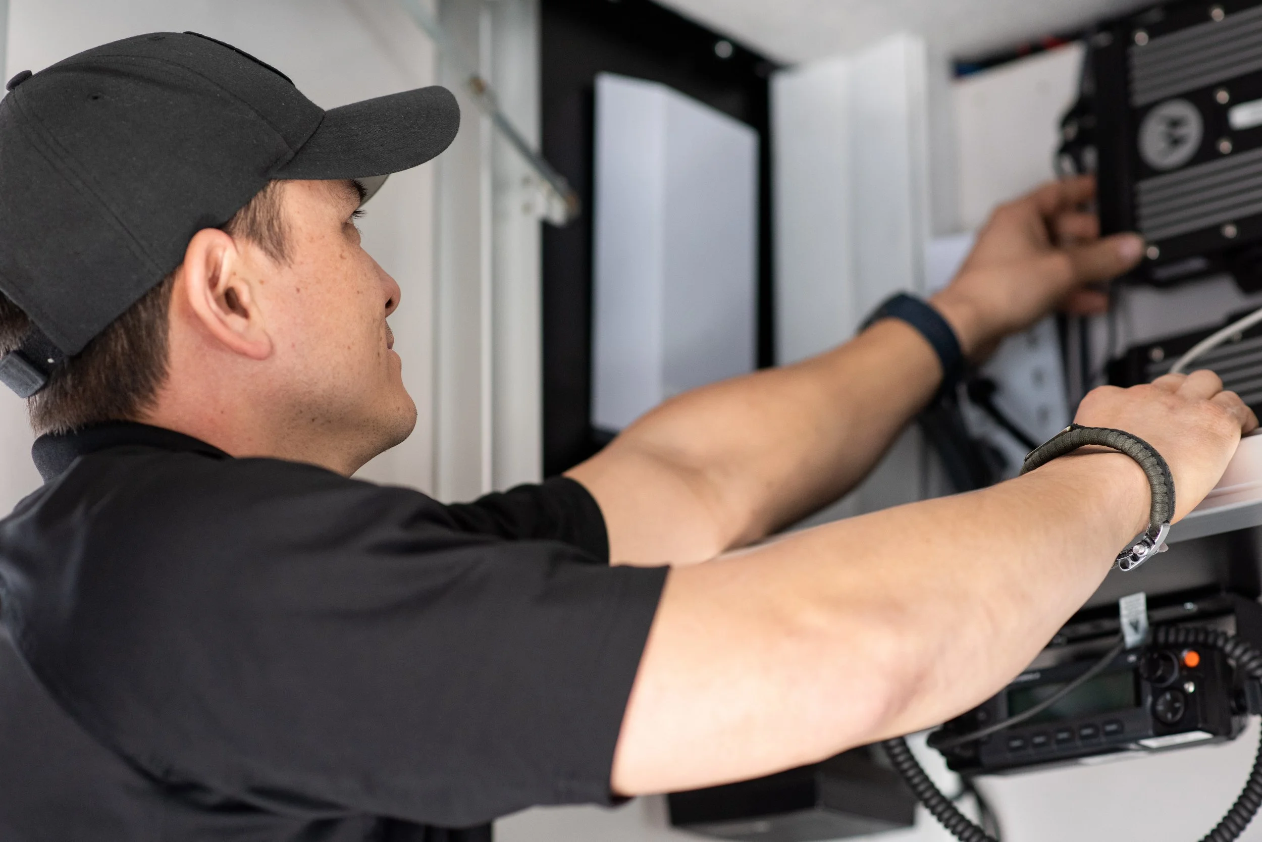 A man wearing a black hat and a black baseball hat is facing away and reaching up to work on some radio equipment in a cabinet.