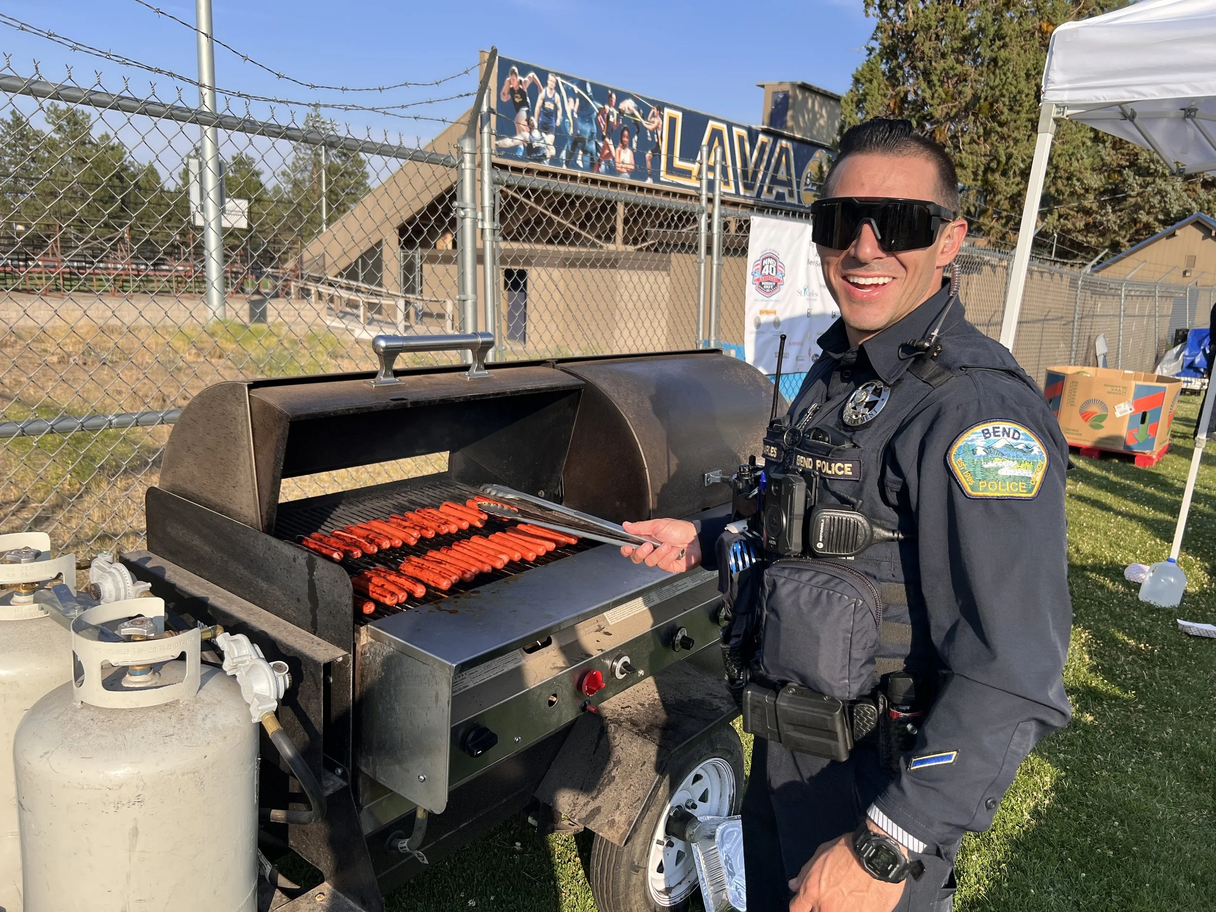 A police officer in uniform stands next to a barbecue grill cooking hot dogs, smiling and holding tongs outside in daylight.