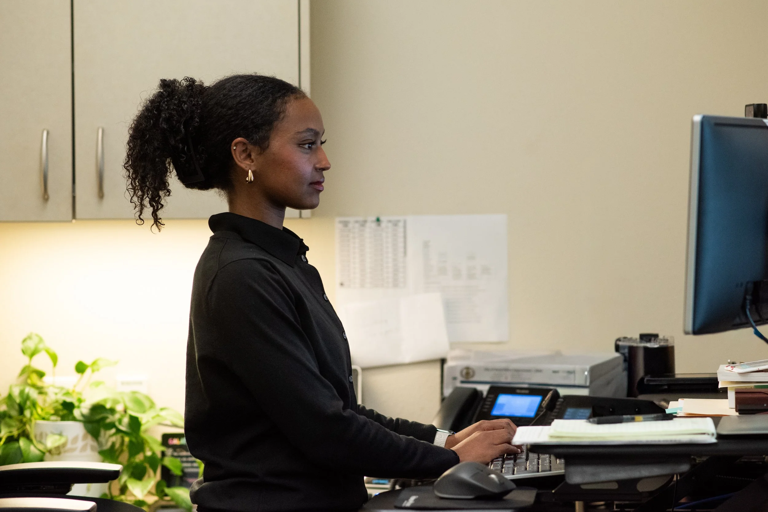 A woman is standing in an office working at a computer station. She is surrounded by storage cabinets on the wall, a phone, paperwork and a plant.