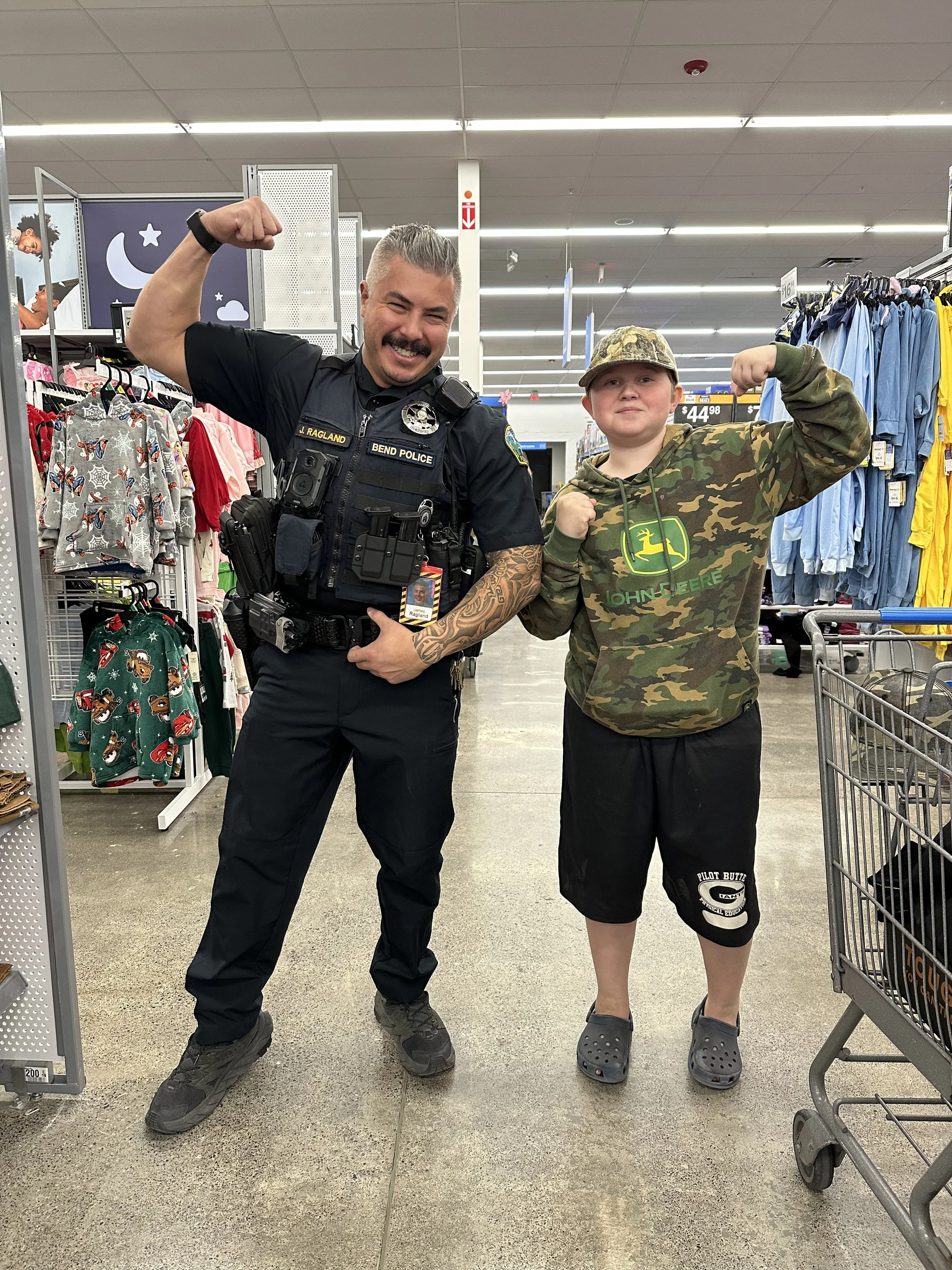 A police officer and a young boy flexing their biceps and smiling inside a store aisle, surrounded by clothing and shopping carts.