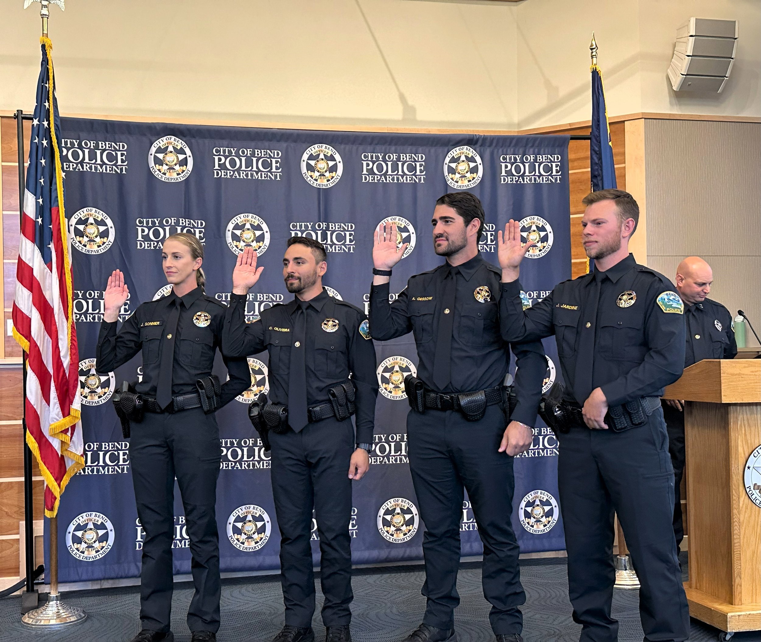 Five police officers standing with their right hands raised during a swearing-in ceremony at the City of Bend Police Department, with an American flag and police department backdrop behind them.
