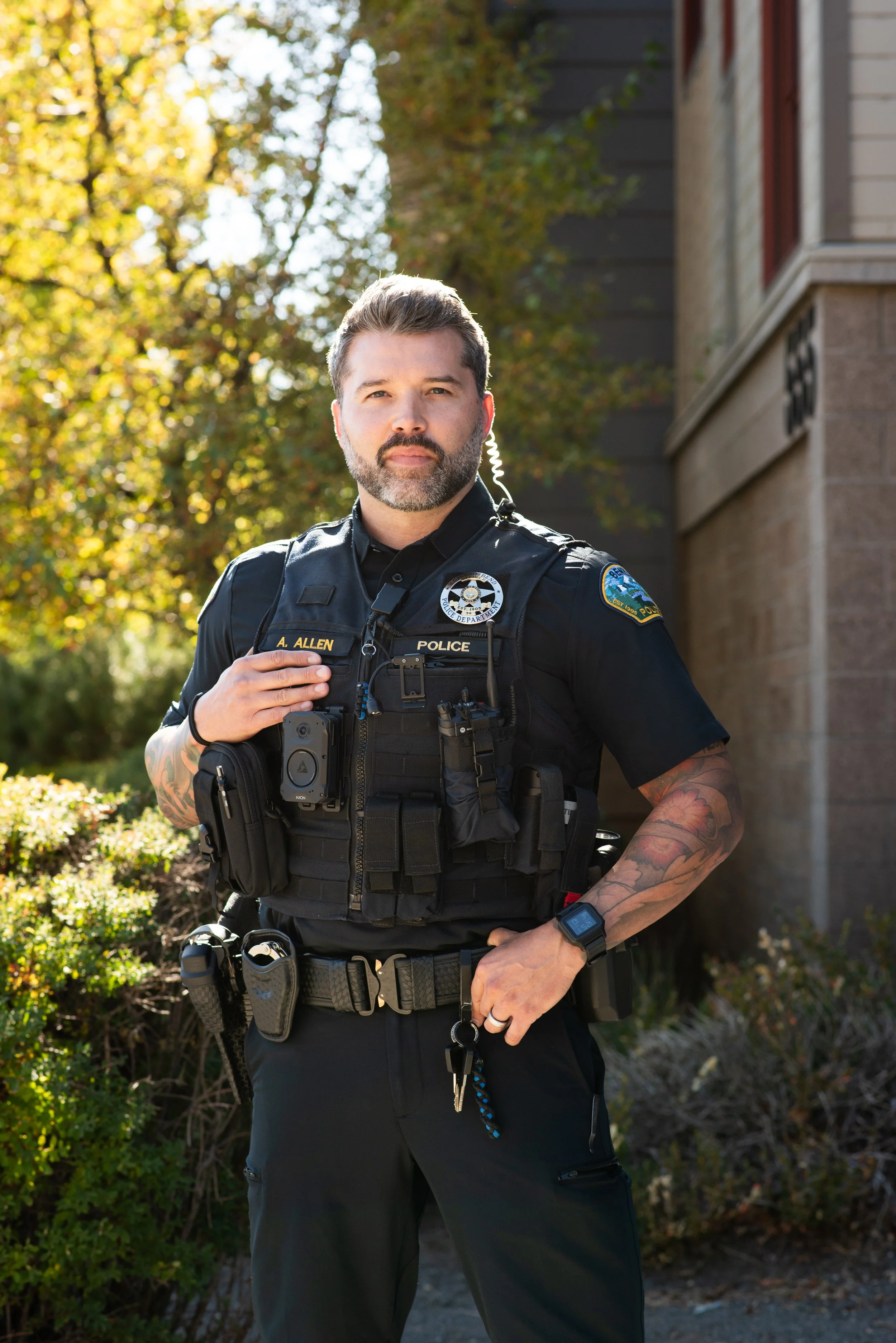 A police officer stands outdoors with a serious expression. He is wearing a black uniform with various tactical gear, including a radio, handcuffs, and pouches. A building and trees with autumn foliage are in the background.