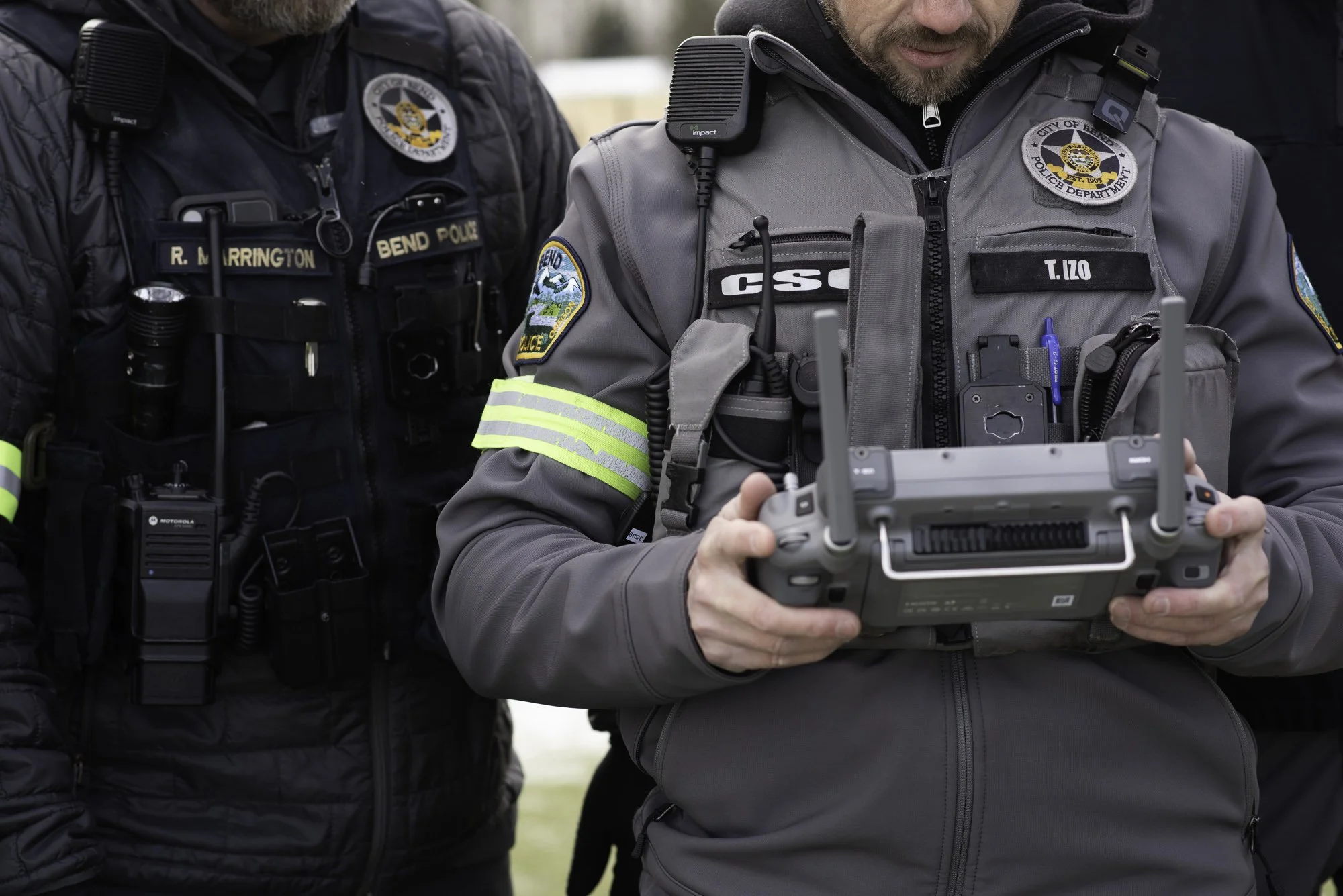 One police officer stands next to a community service officer holding a drone remote control, stand outdoors in uniform with badges and equipment.