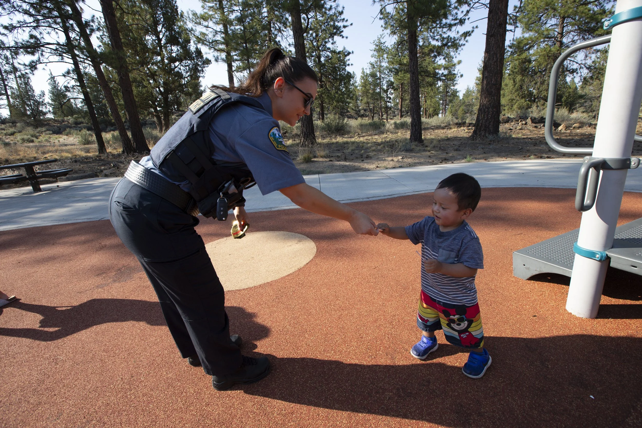 A female community service officer in uniform and sunglasses is giving a small boy a high five at a playground, with a background of trees and a blue sky.