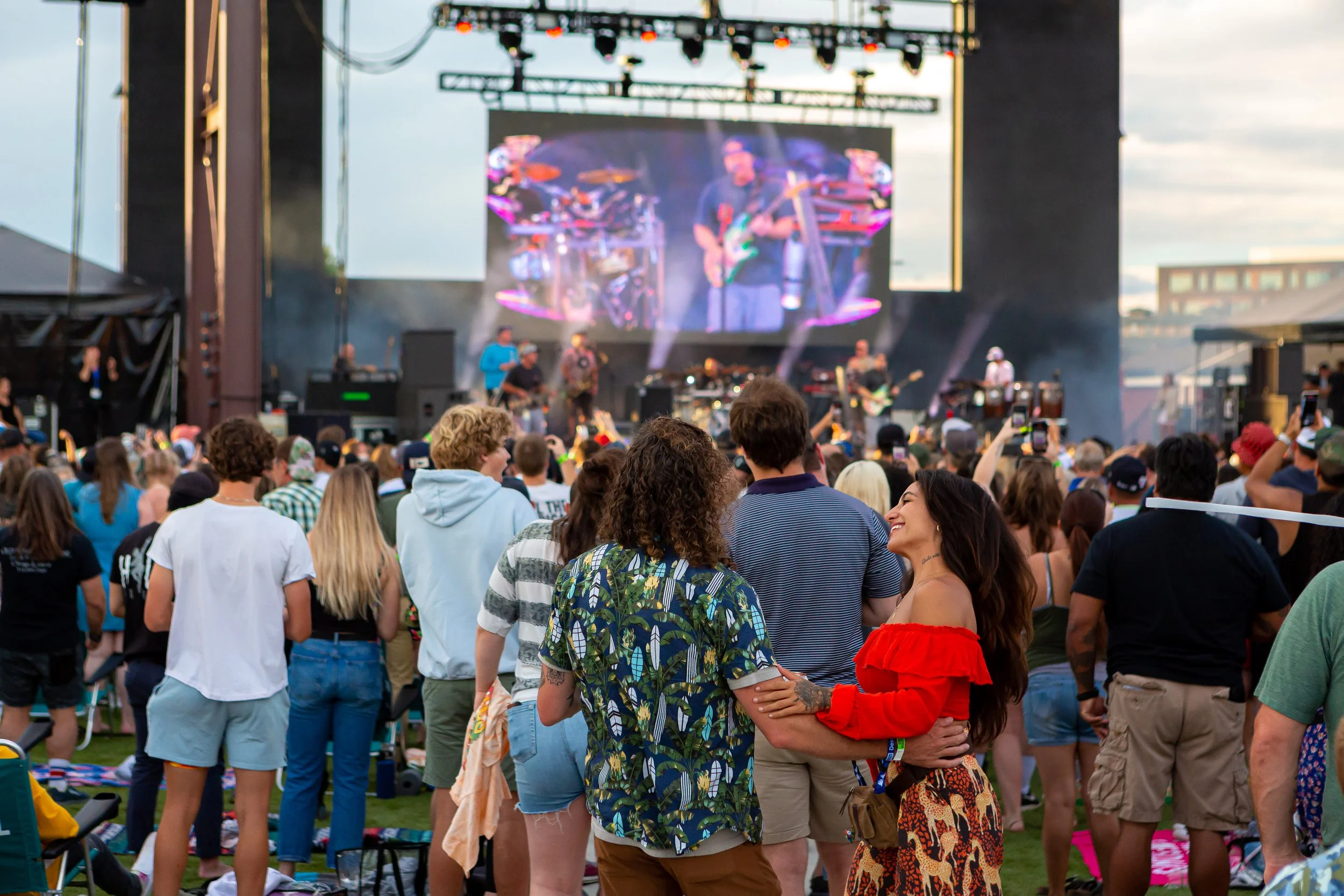 A large group of people attend a rock concert outdoors. They have their backs to the camera and are watching the performers on stage. A large screen shows the performers. A woman in a red shirt smiles at the person next to her.