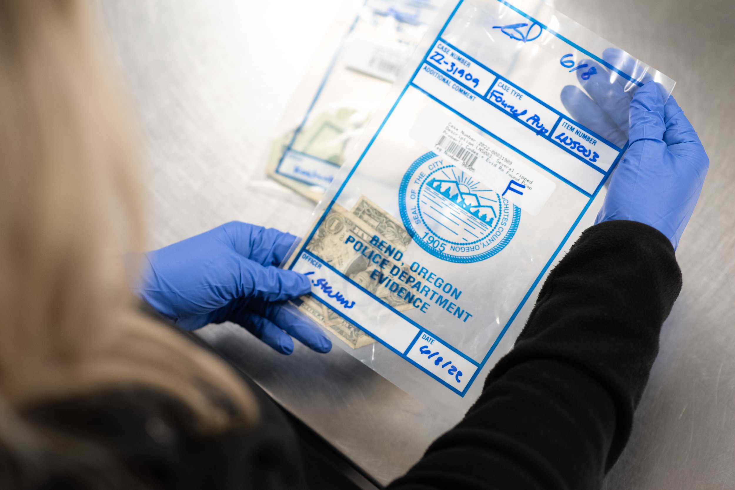 A person wearing blue disposable gloves is holding a clear plastic evidence bag containing torn pieces of paper money.