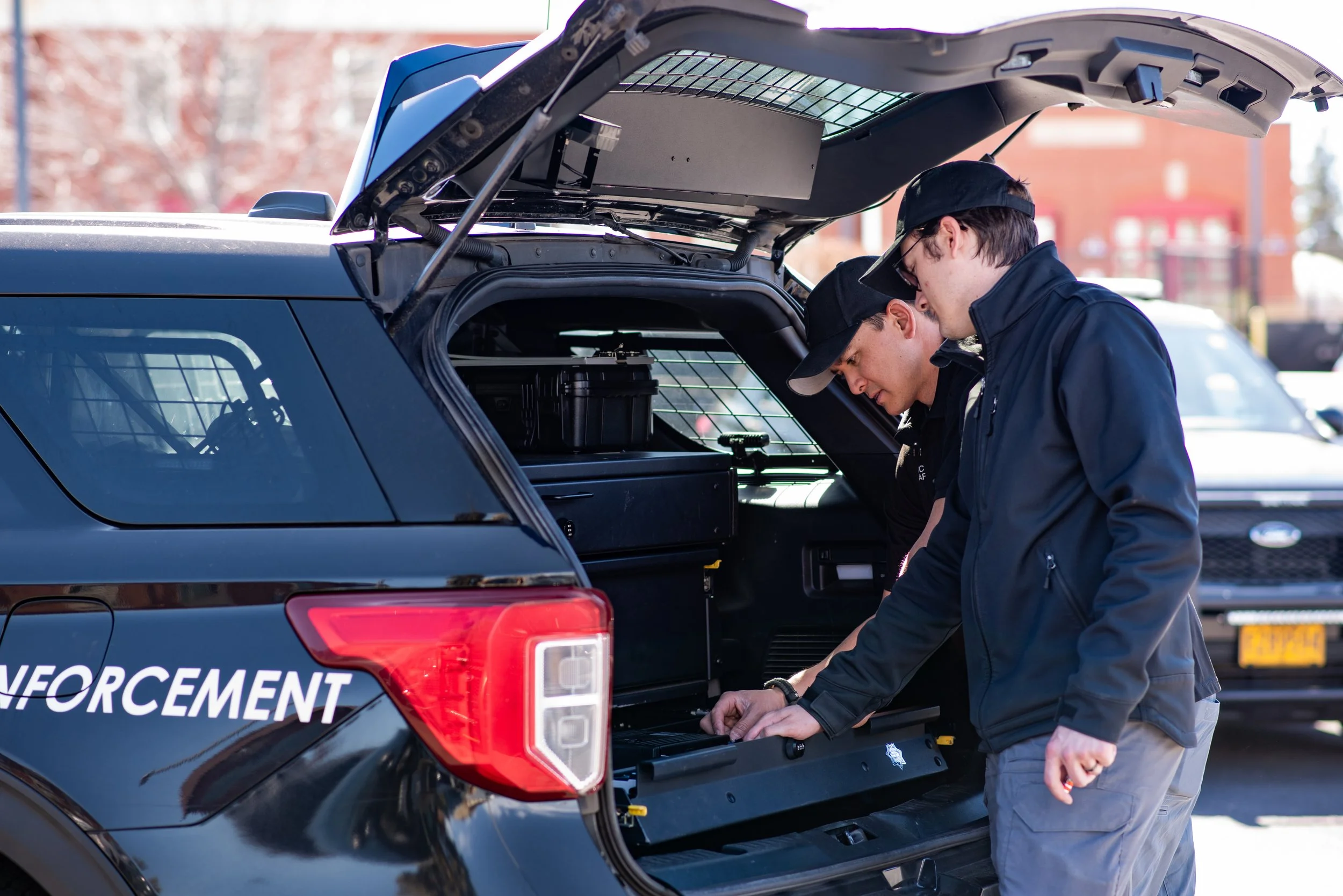 Two men are outside at the black of a black police SUV. They have the rear hatch open and are looking inside to work on radio equipment in the cargo area of the vehicle. The men are both wearing black clothing.