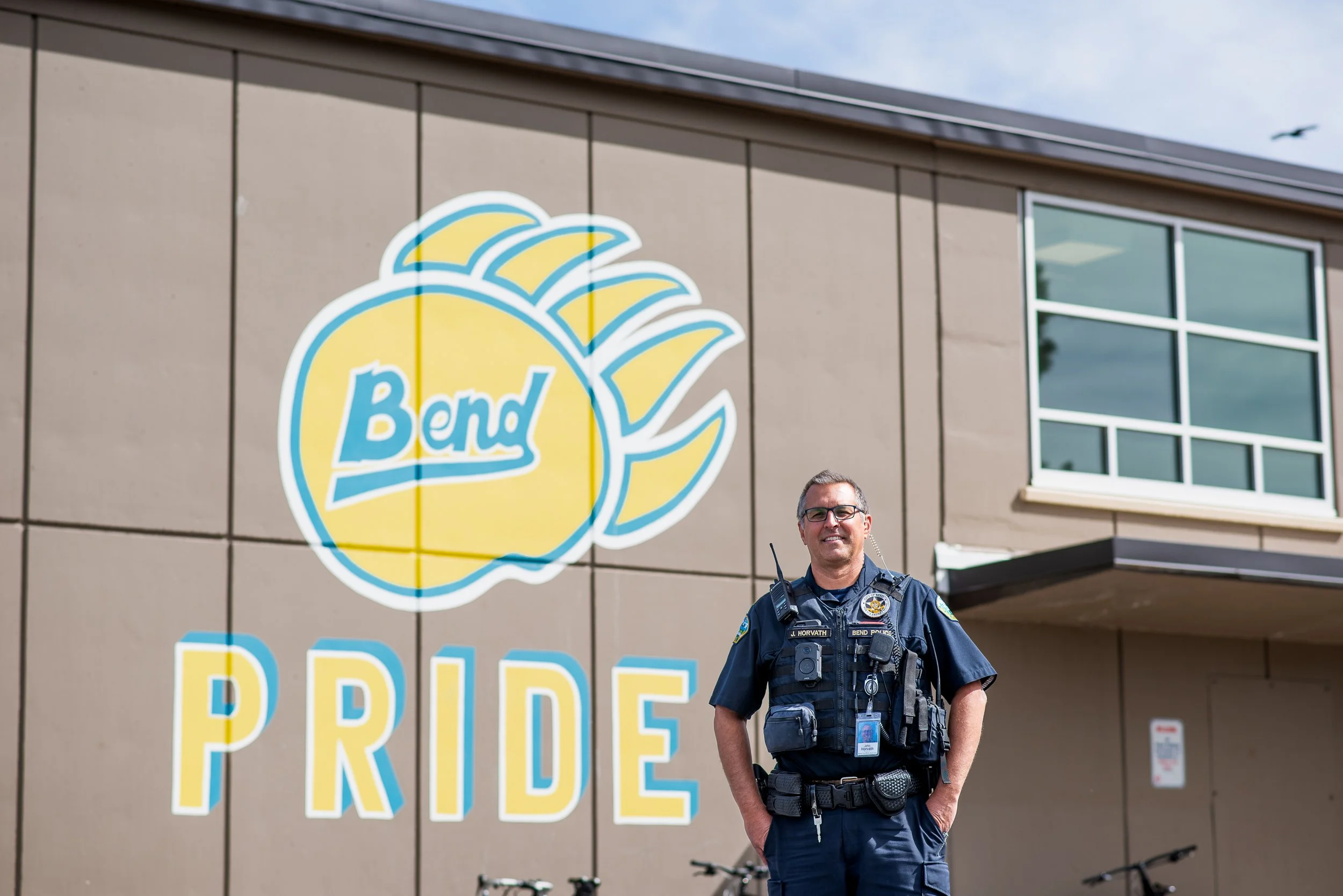 A police officer standing outside a building with a large mural of a yellow beach ball and the words "Bend PRIDE" on it.