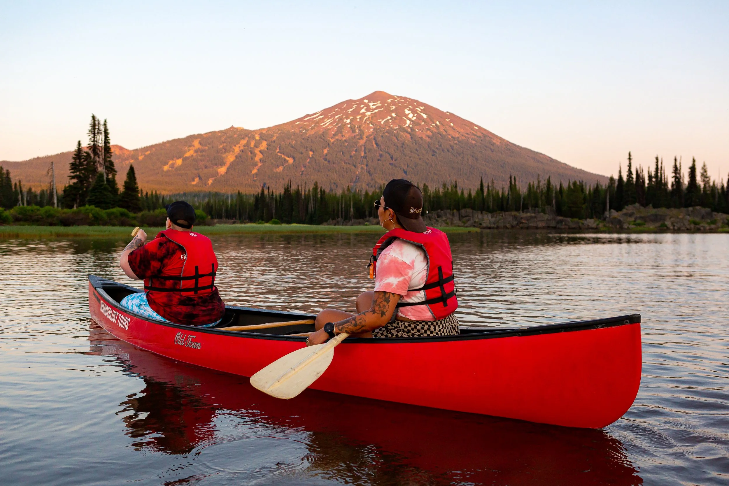Two people in red life vests sit in a red canoe and paddle on a lake with a mountain and trees in the distance.