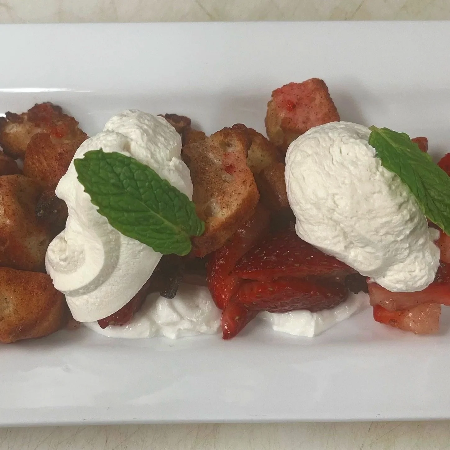Fried food with whipped cream, strawberries, and mint leaves on a white rectangular plate.