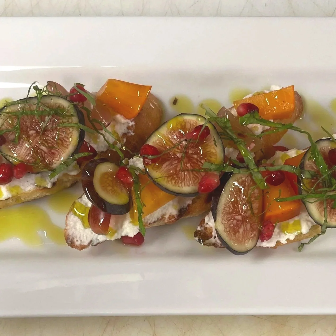 Canapés with figs, cherry tomatoes, topped with pomegranate seeds and herbs, drizzled with olive oil on a white rectangular plate.