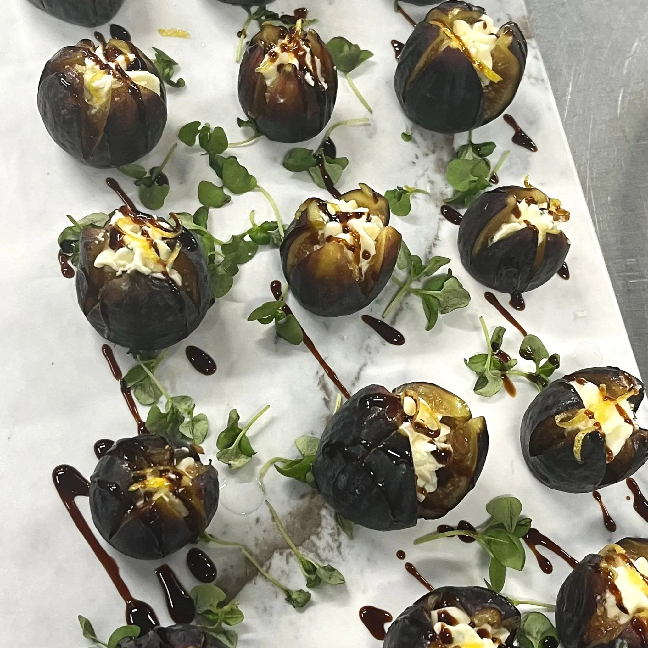 Stuffed figs drizzled with balsamic glaze, garnished with microgreens and cream, arranged on a white surface.