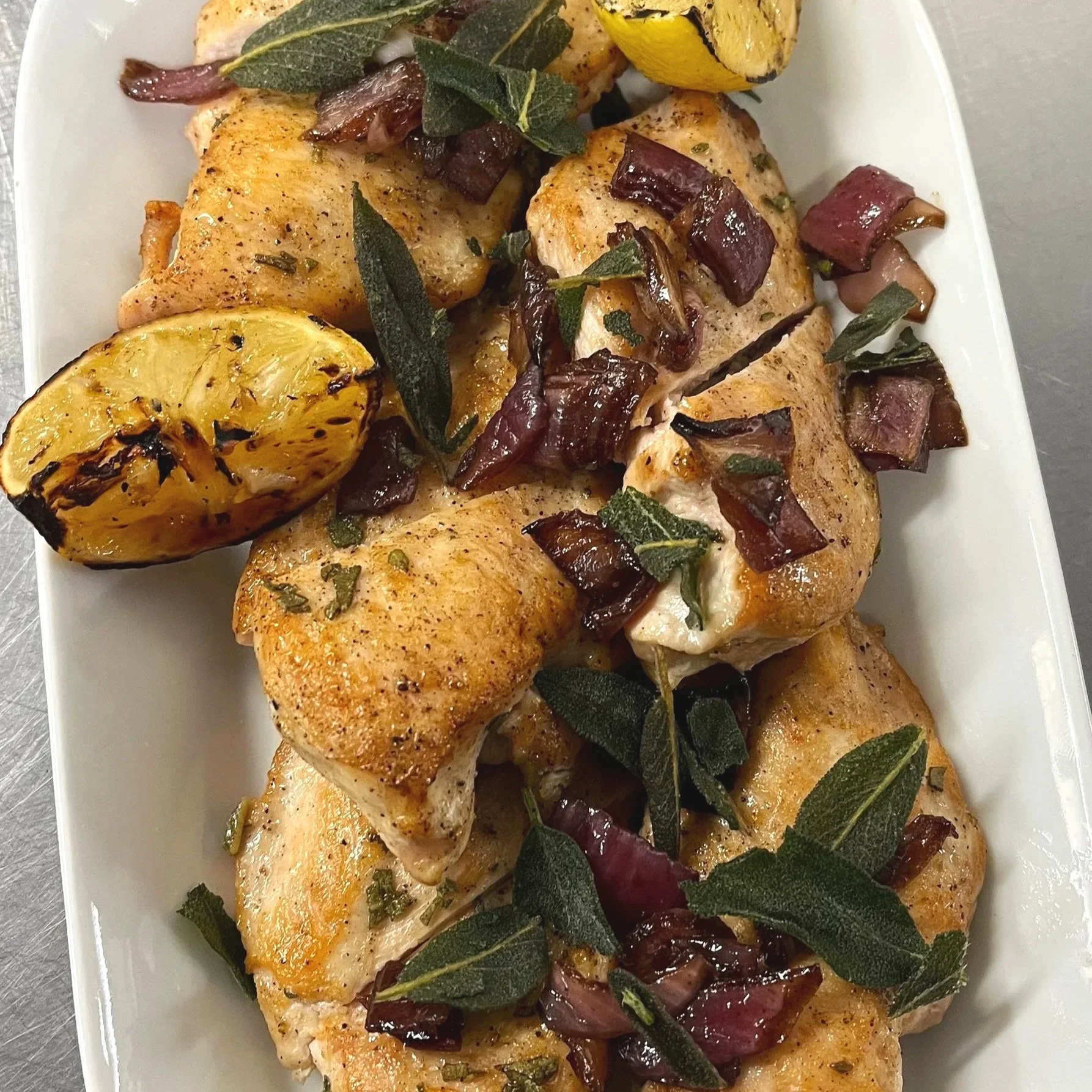 Grilled chicken pieces garnished with roasted lemon halves, chopped shallots, and fresh sage leaves on a white serving platter.