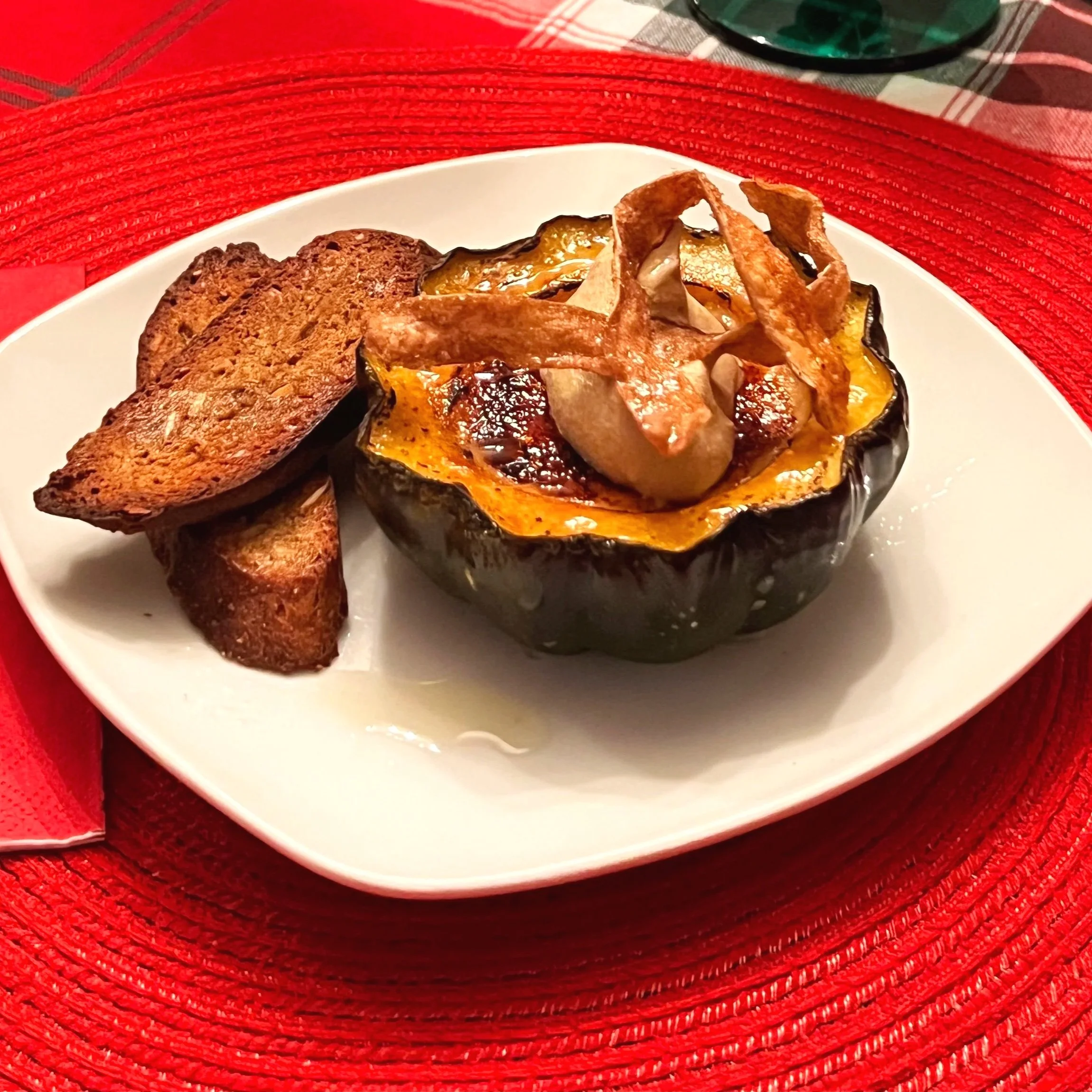 Stuffed acorn squash topped with bacon strips, served with toasted bread slices on a white plate, placed on a red placemat.