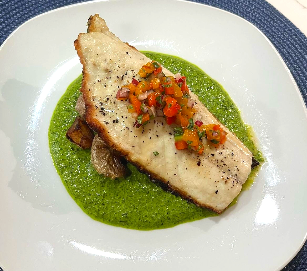 A plated serving of grilled fish topped with a colorful chop of diced vegetables, resting on a bed of green sauce on a white dish.