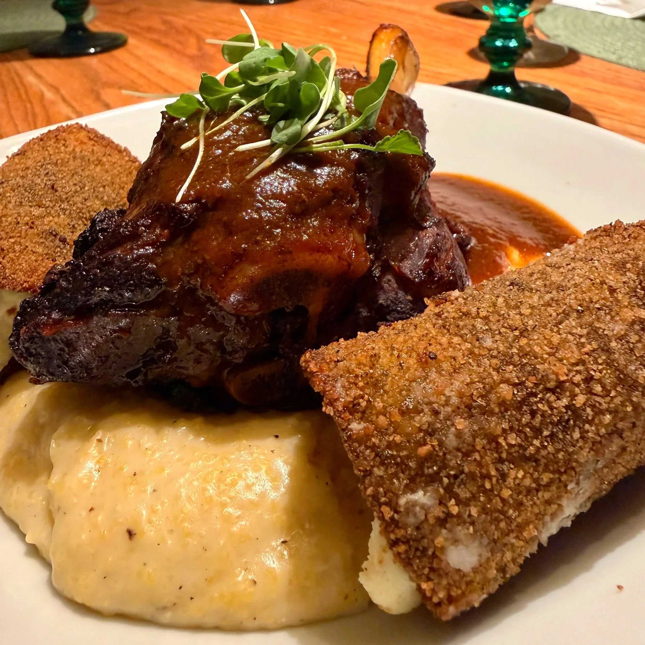 A plate of food with a braised piece of meat with sauce and microgreens on top, two breaded and fried items, and a serving of mashed potatoes, on a wooden table.