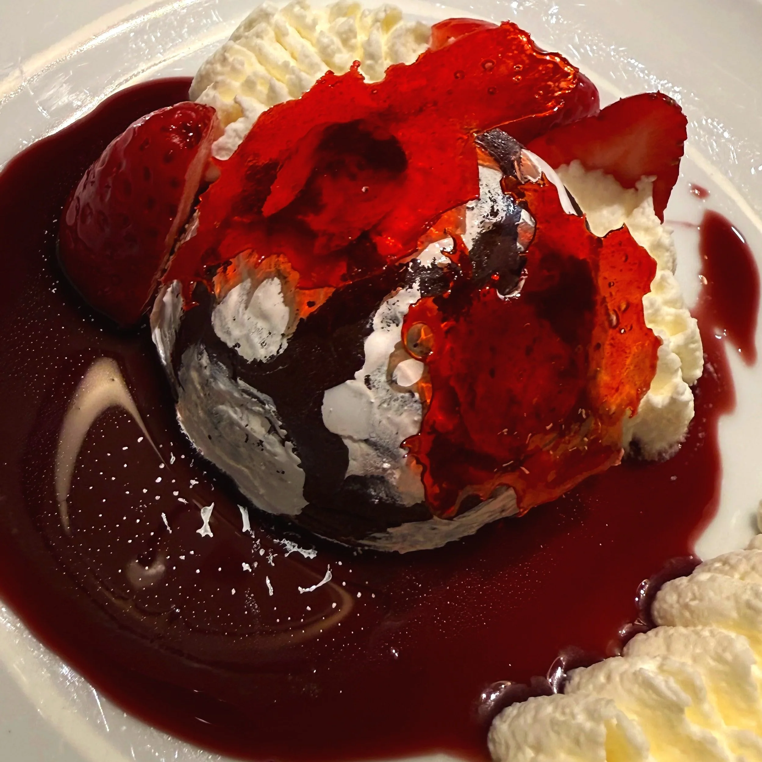 A dessert featuring a scoop of vanilla ice cream topped with red fruit sauce, surrounded by whipped cream and additional red fruit syrup.