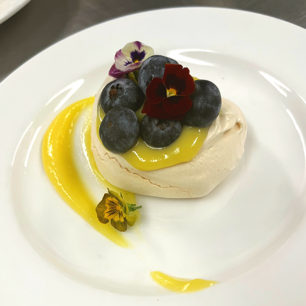 A dessert on a white plate with fresh blueberries, edible flowers, lemon curd, and a small dollop of cream or mousse.