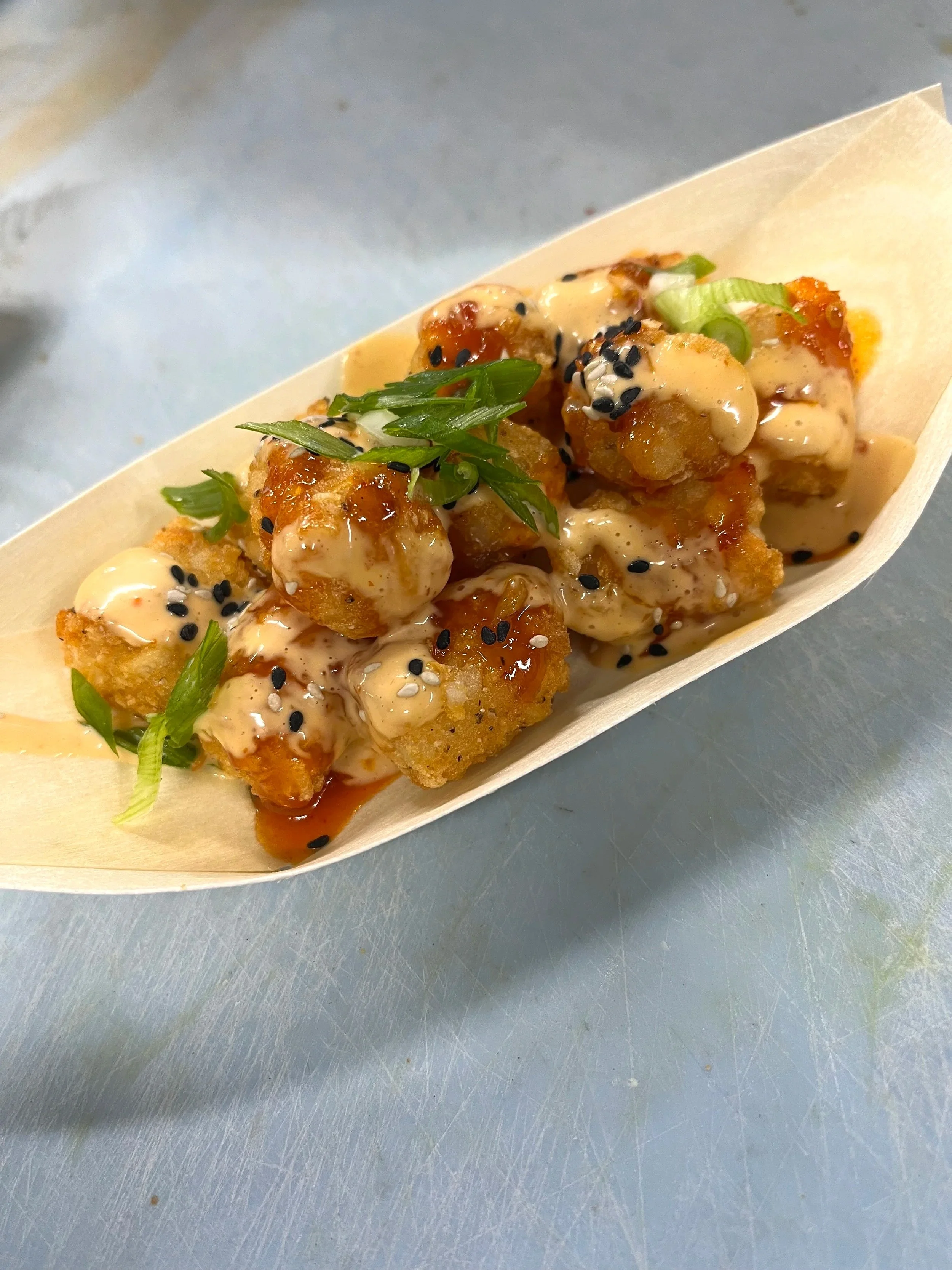 Fried pieces of tater tots topped with sauce, sesame seeds, and chopped green onions in a paper tray.