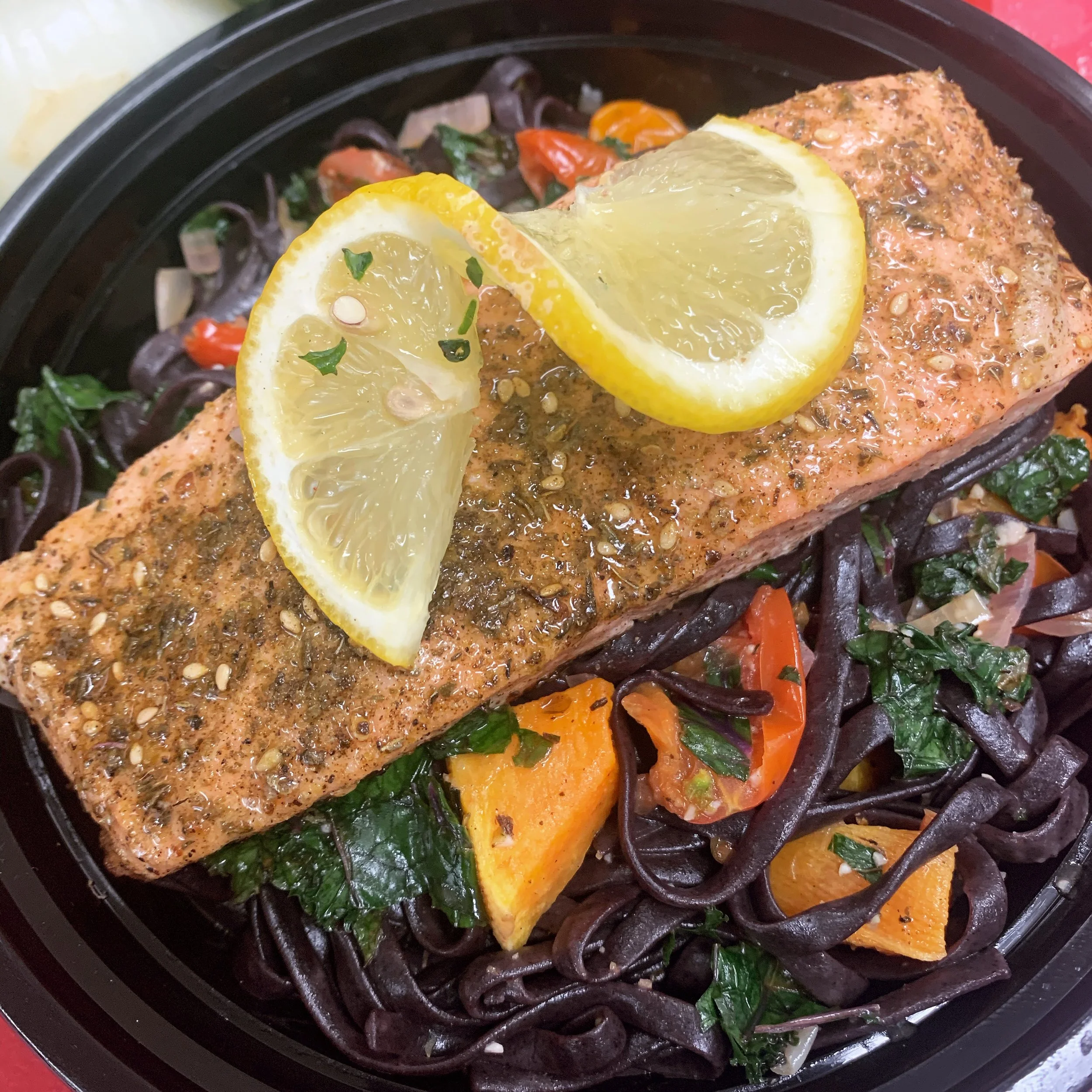 Cooked salmon fillet topped with lemon slices and herbs, served over black pasta with roasted tomatoes, sweet potatoes, and kale.
