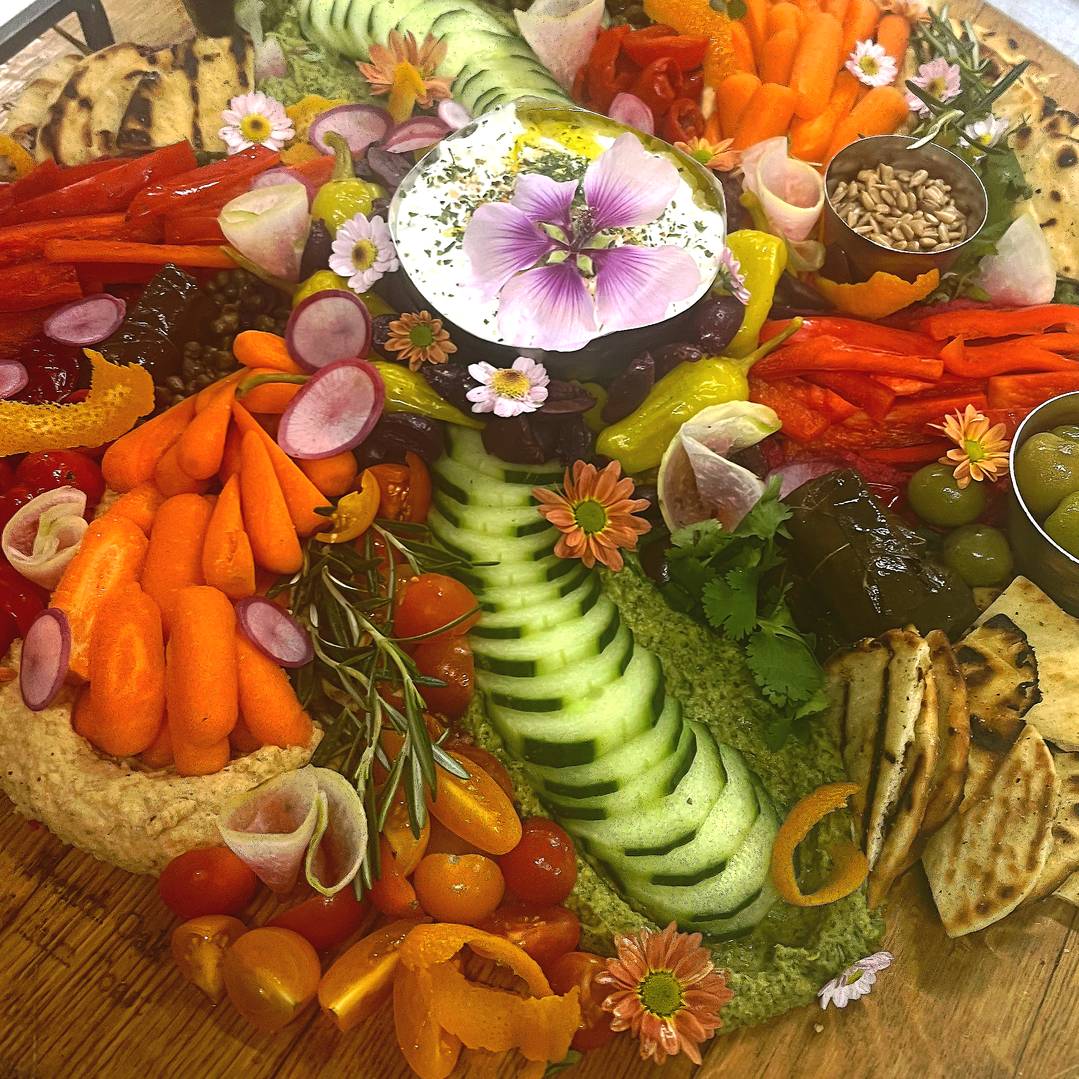 A colorful vegetable platter with cherry tomatoes, cucumbers, bell peppers, carrots, radishes, grilled bread, sunflower seeds, herbs, and decorative flowers arranged around a bowl of dipping sauce.