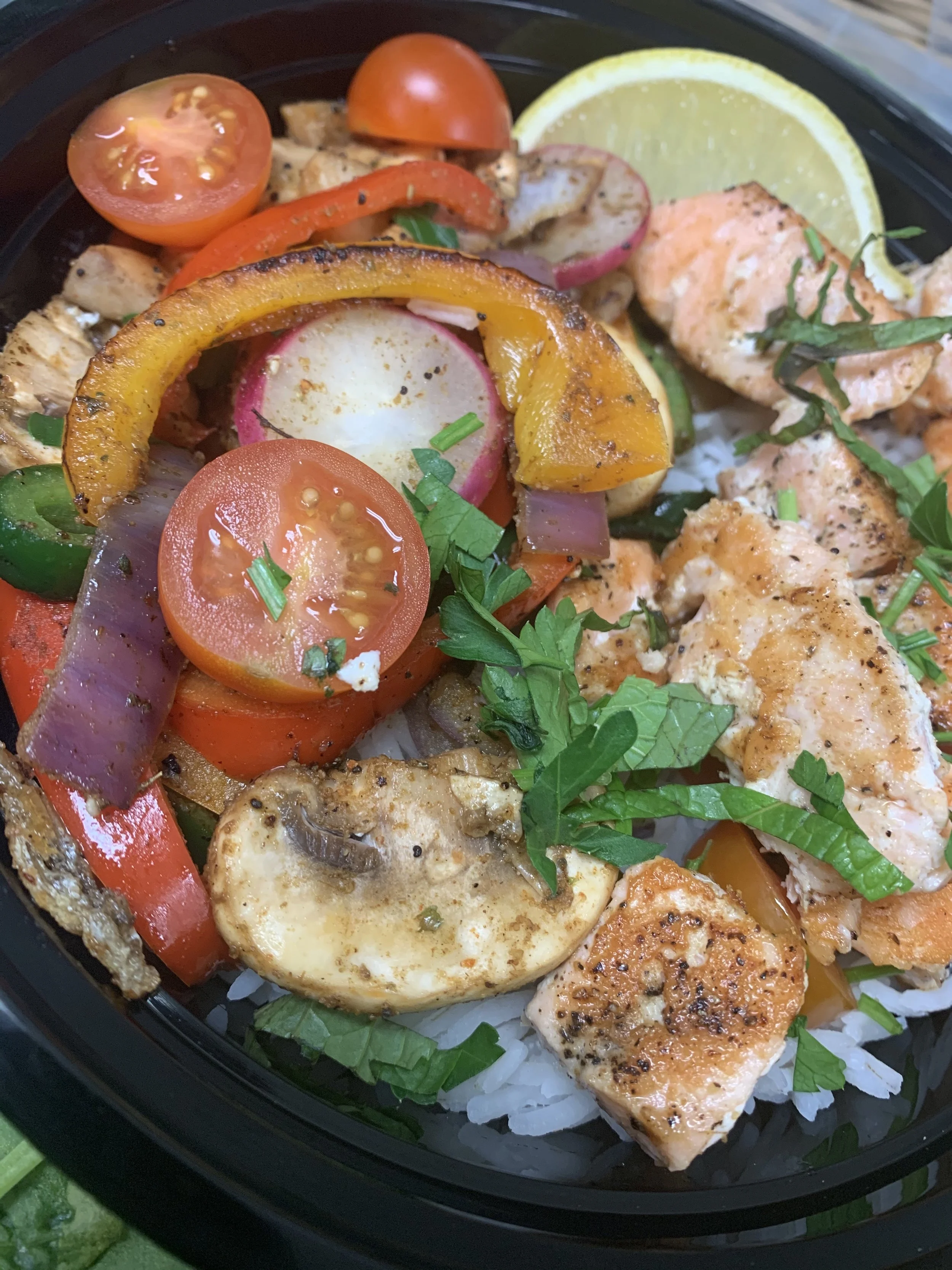 Grilled chicken and vegetable rice bowl with cherry tomatoes, sliced radishes, bell peppers, mushrooms, and garnished with fresh herbs, with lemon slices on the side.