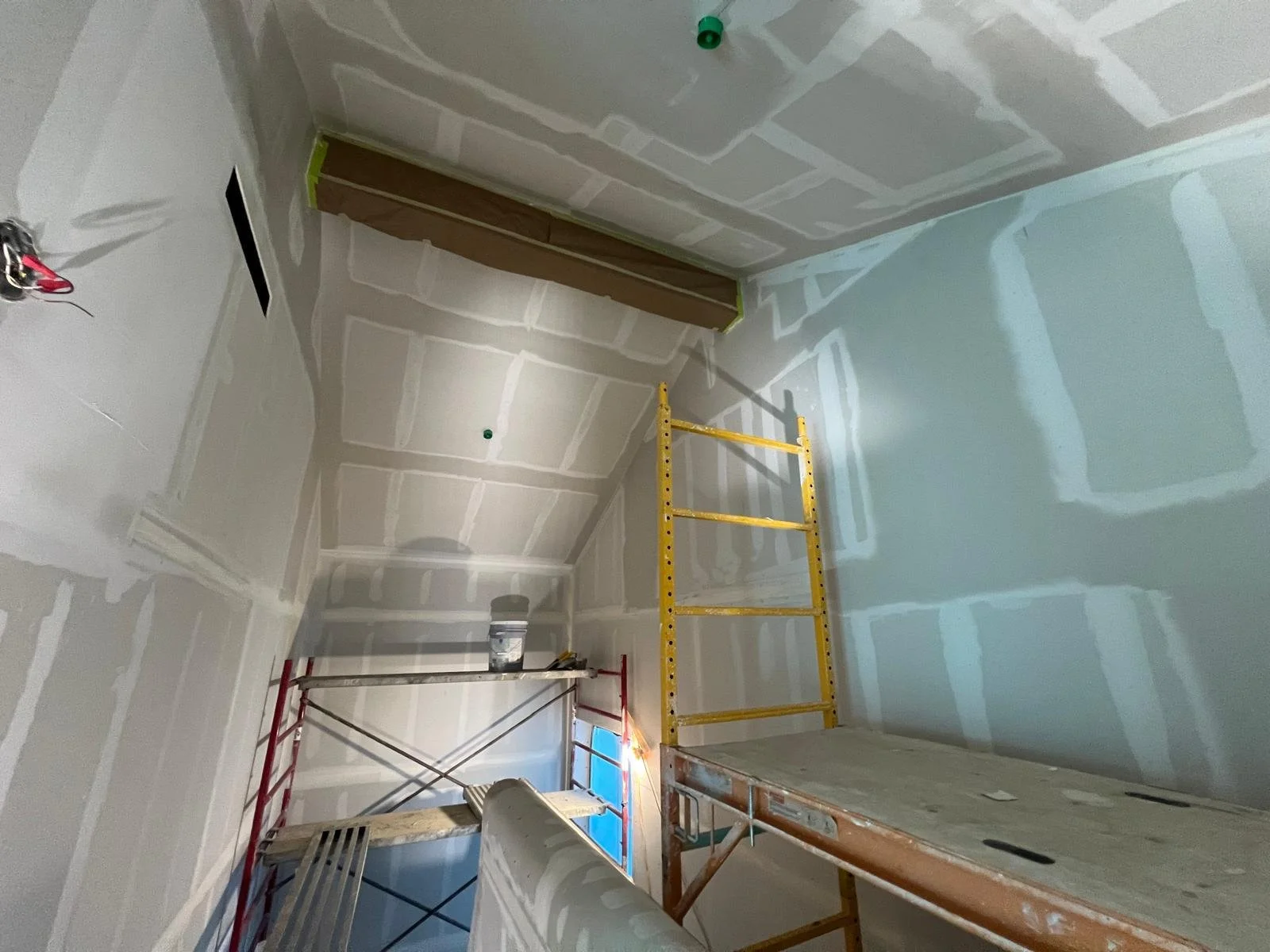 Room under construction with drywall installed, a yellow ladder, and a scaffold with tools. The ceiling and walls are partially finished with visible joint compound.