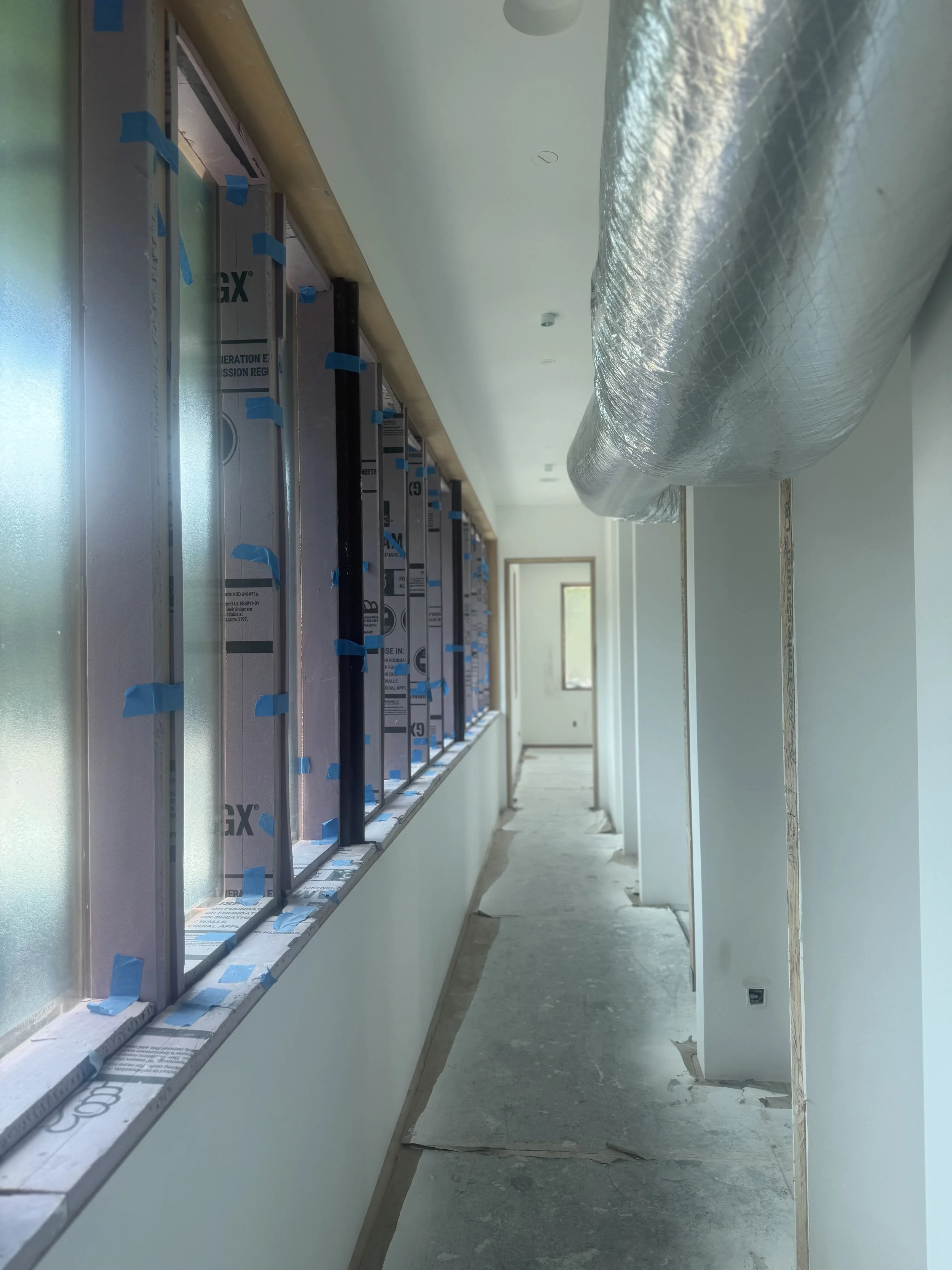 Interior of a building under construction with a long hallway, windows on the left, and a large duct running along the ceiling.