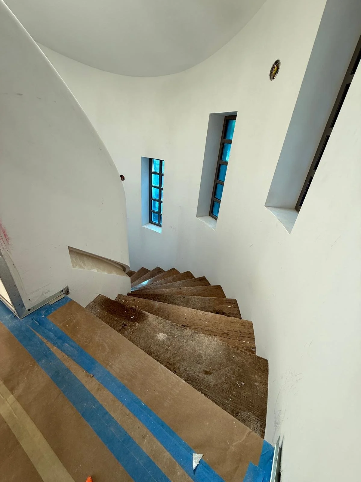 Interior view of a staircase under construction with wooden steps, three narrow windows with blue glass, white curved wall, and protective blue tape on the floor.