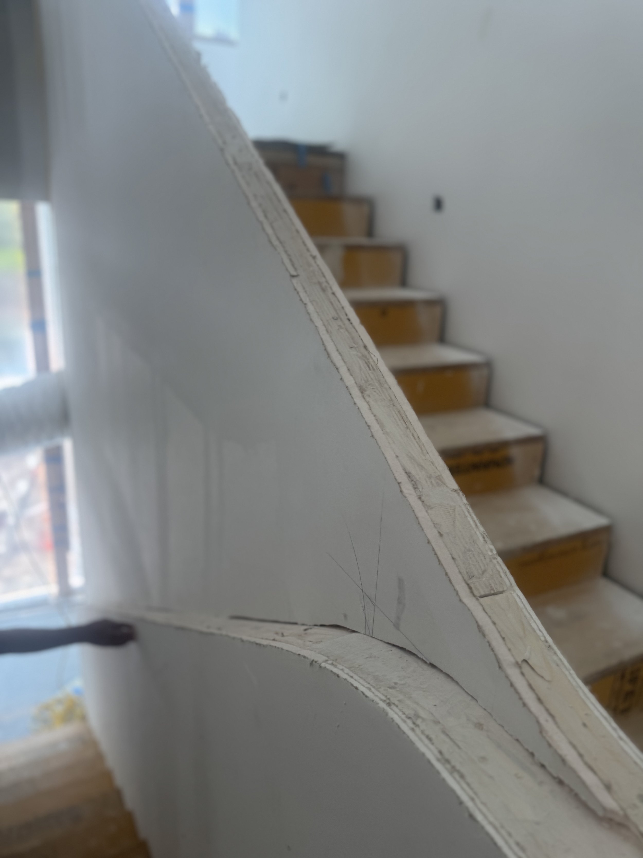 Close-up of a white painted stair banister with exposed wood underneath in a house under construction or renovation, with a staircase in the background.