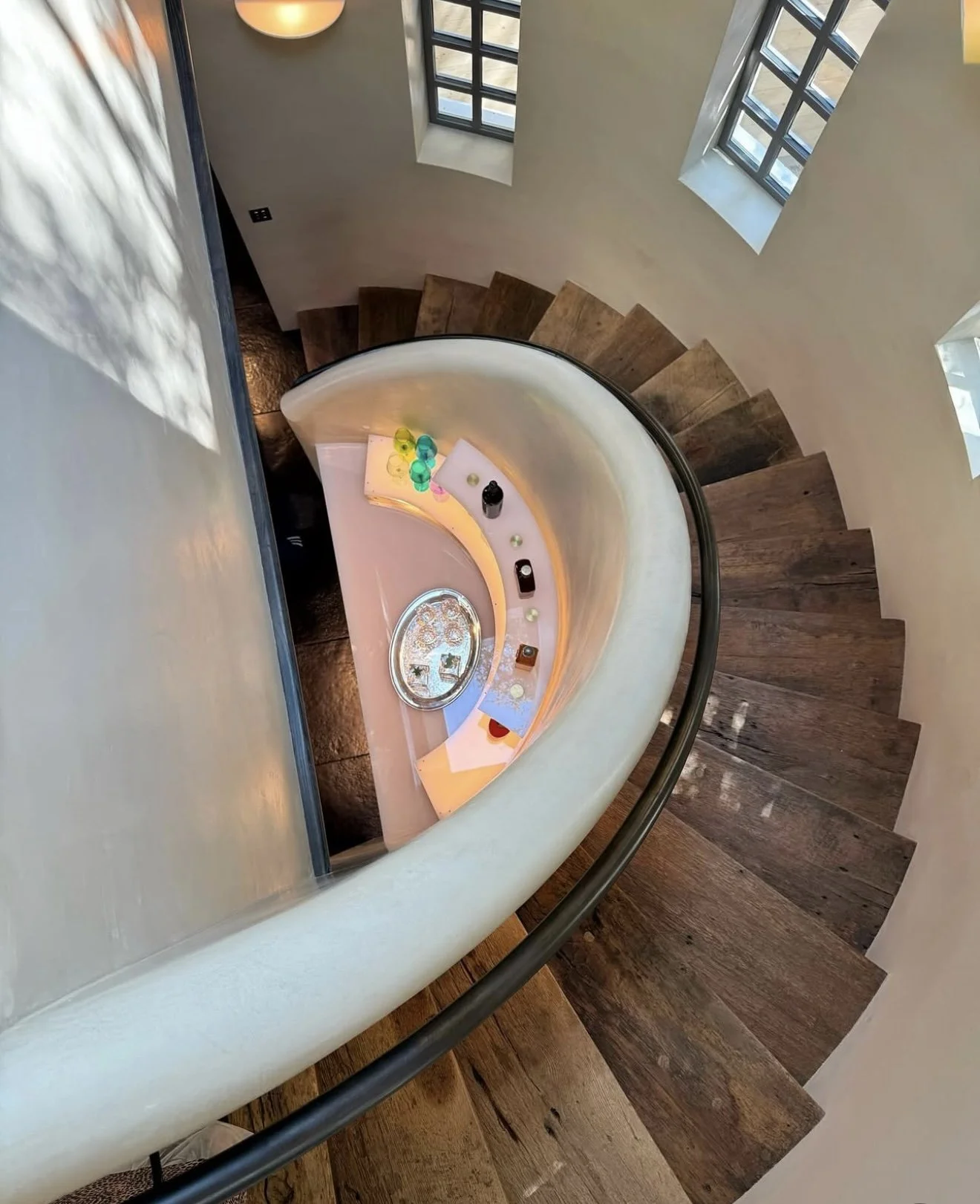 A spiral staircase with wooden steps and a white curved railing. At the bottom of the staircase, there is a small landing with decorative items, including colorful balls and a silver tray, viewed from above near a window with sunlight casting shadows.