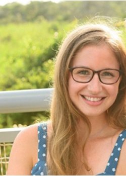 Smiling woman with curly hair, wearing glasses and hoop earrings, in a casual office or cafe setting.