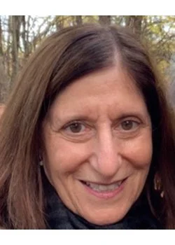 Close-up portrait of a woman with dark hair, wearing clear glasses, a nose ring, and a dark green sweater, looking thoughtfully to the side indoors.