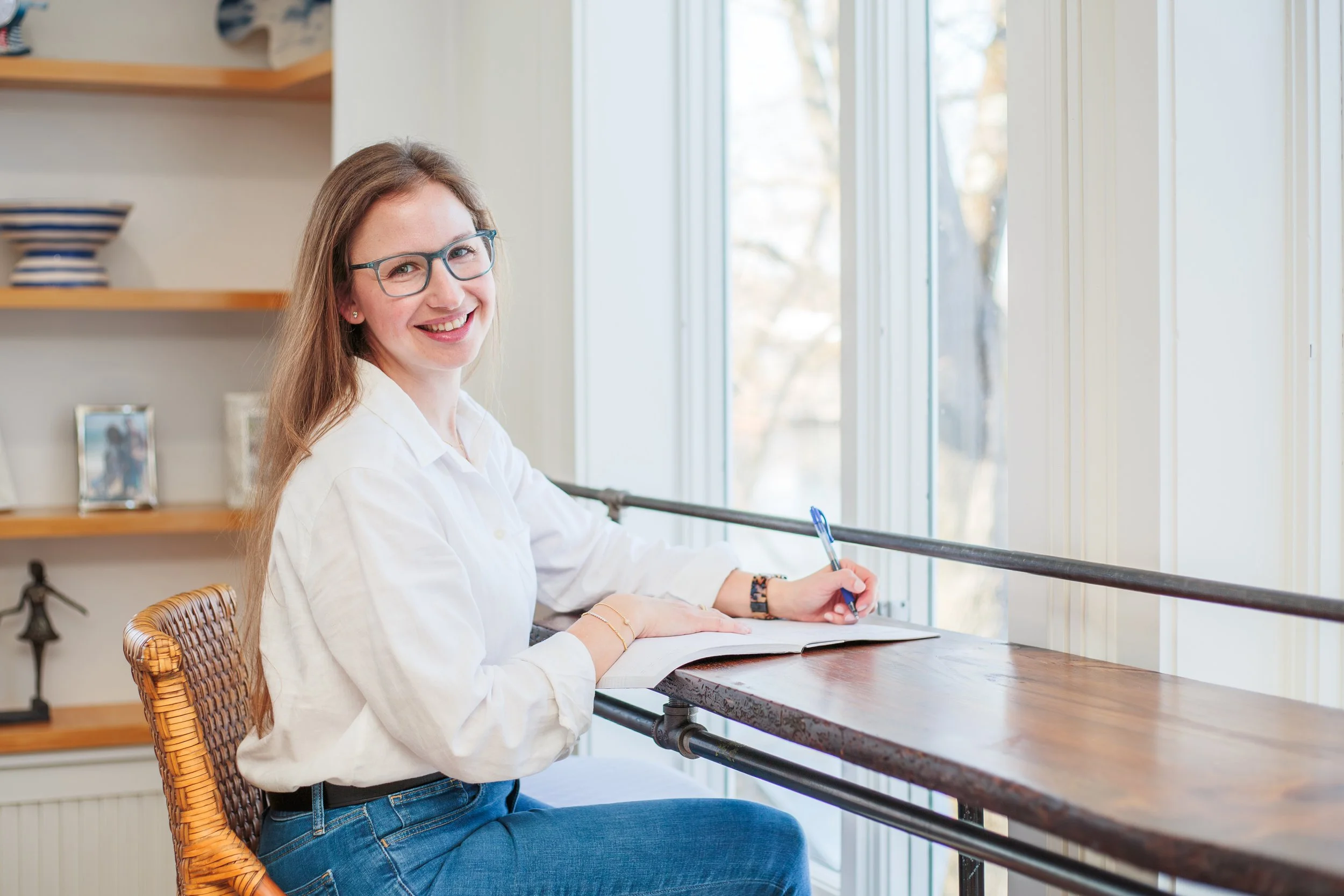 Smiling woman with curly hair, wearing glasses and hoop earrings, in a casual office or cafe setting.