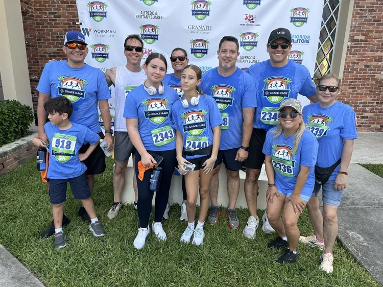 Families and runners in Grace Race 5K shirts smiling in front of the sponsor banner.