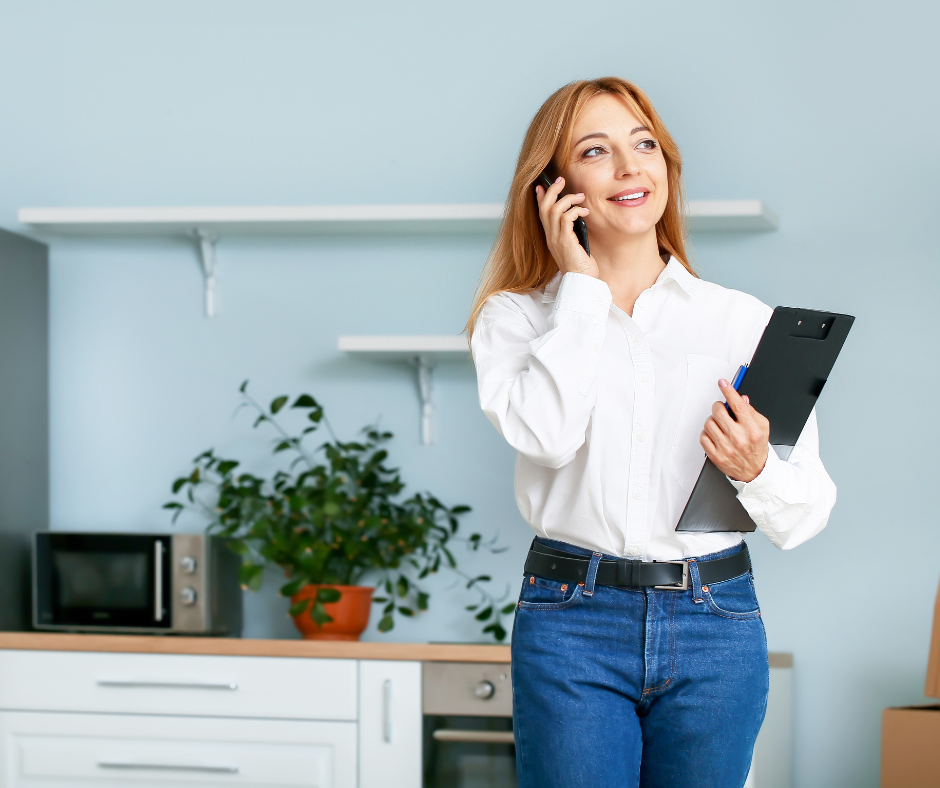 A woman with red hair smiling and talking on her cellphone while holding a folder in her other hand, standing in a modern kitchen with plants, microwave, and shelves.