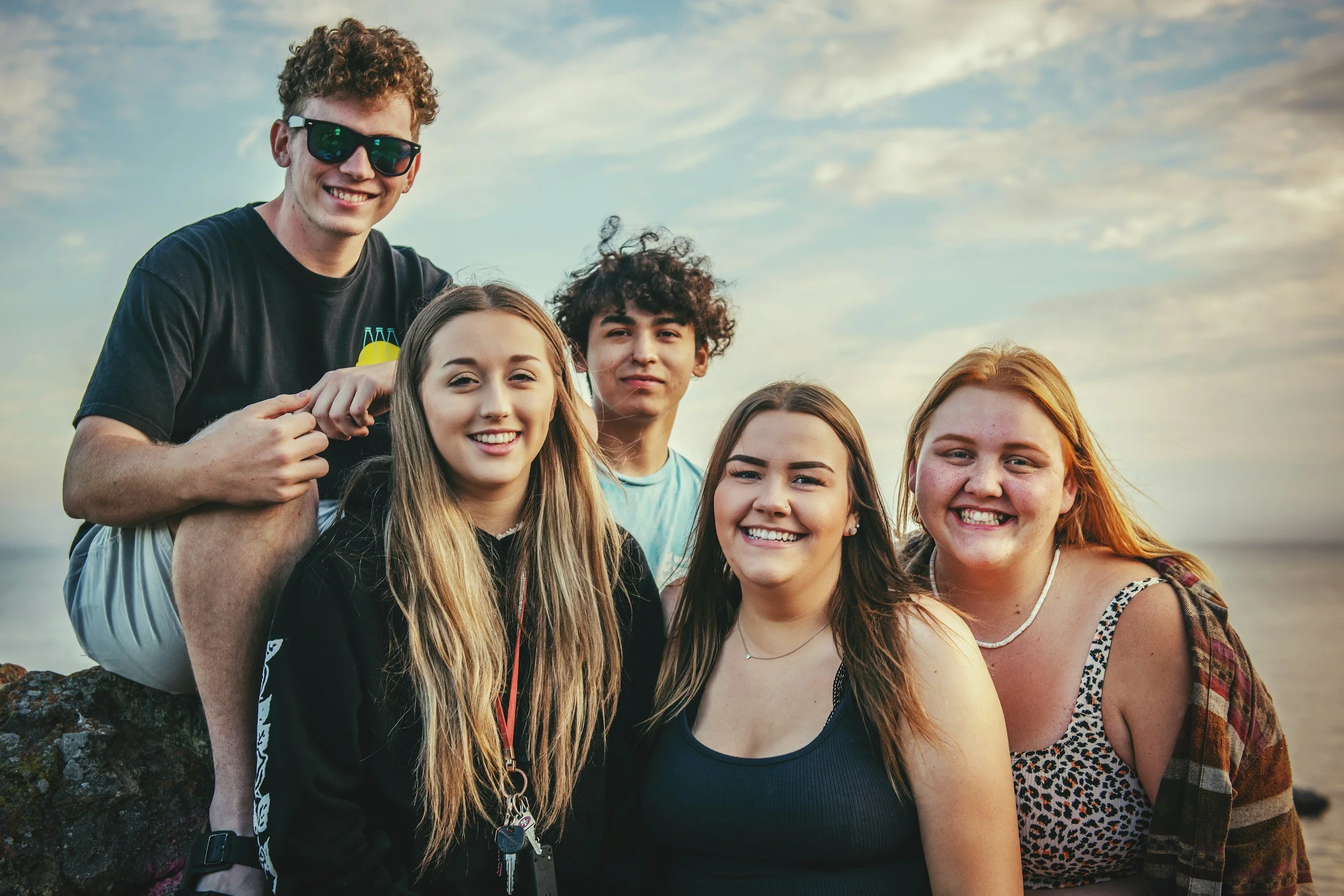 Group of five teenagers smiling together outdoors near the ocean during sunset.