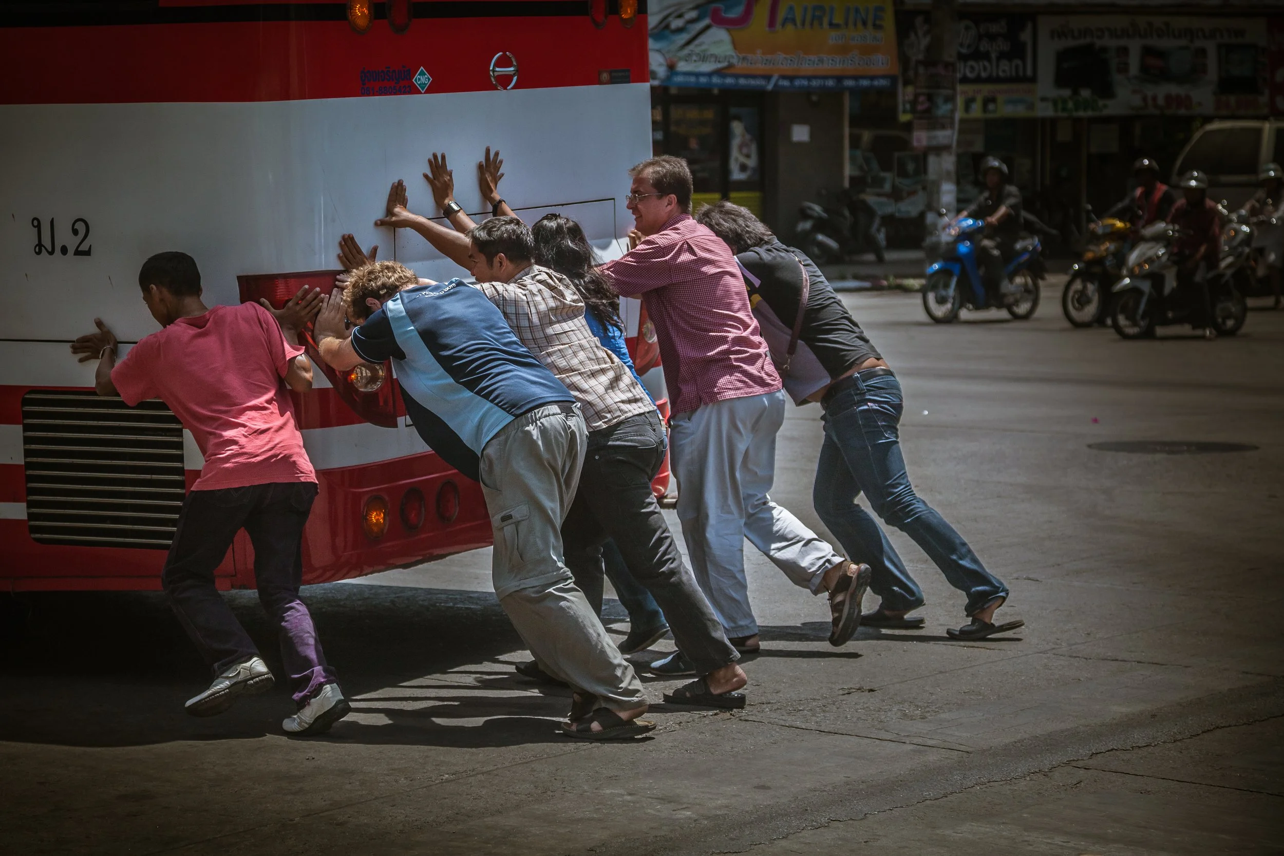 Group of people pushing a large red and white bus from the back on a street in an urban area, with motorbikes and shops in the background.